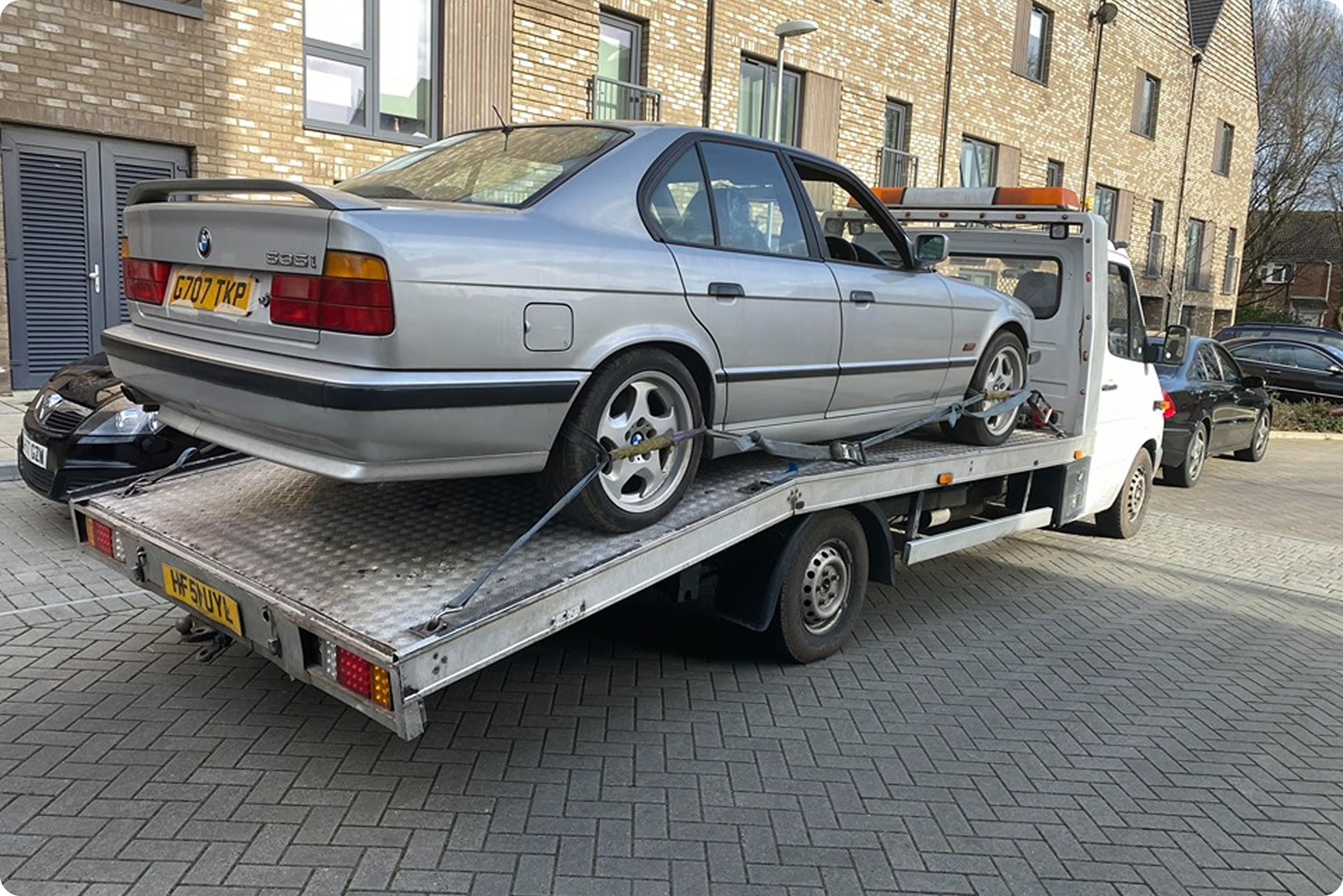 A silver BMW E30 3 Series car on a flatbed tow truck in an urban area with brick apartment buildings in the background.