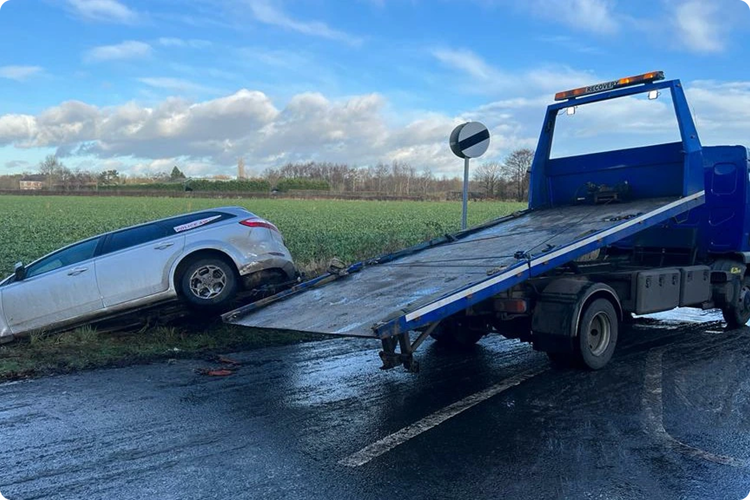 A car transporter truck carrying a silver car on a roadside with a field and blue sky with clouds in the background.