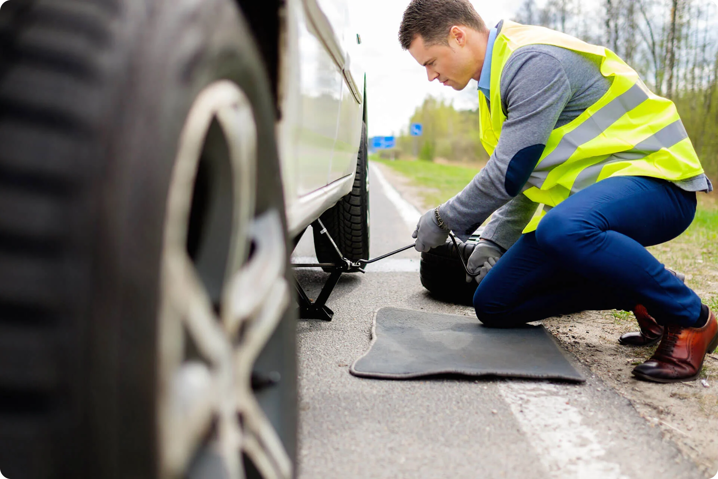A man wearing a yellow safety vest and blue pants is kneeling on the road next to a car, changing a flat tire.