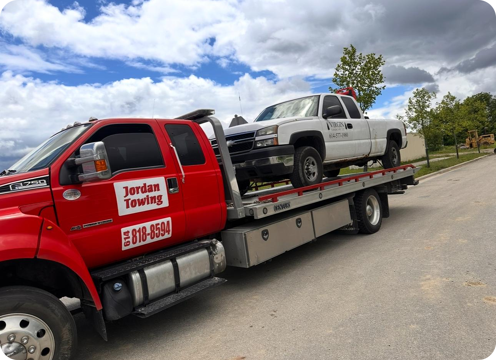 A red tow truck transporting a white pickup truck. The tow truck has a sign that reads "Jordan Towing" with a phone number. The pickup truck has writing on the door that reads "VIRGIN" with a phone number. The background shows a partly cloudy sky, some trees, and a dirt area with construction equipment.