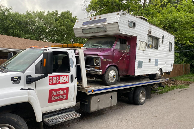 A tow truck carrying a vintage camper van with a maroon front cab and a white living area behind, parked on a residential street with trees and houses in the background.