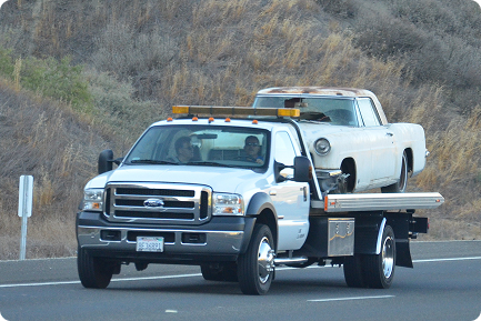 A white tow truck transporting an empty vintage white car on a highway.