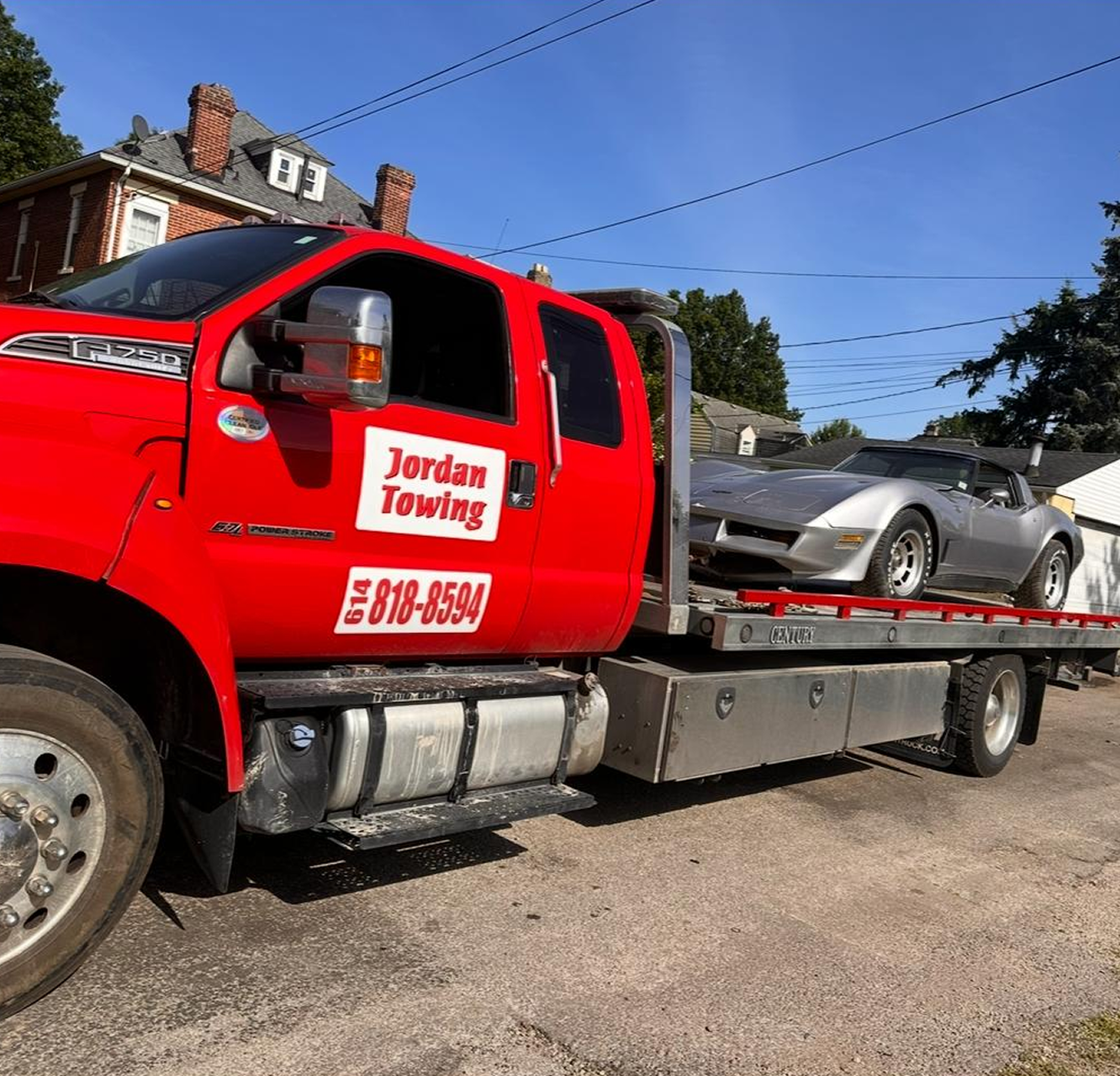 A red tow truck carrying a silver sports car parked on the side of a residential street under a clear blue sky.