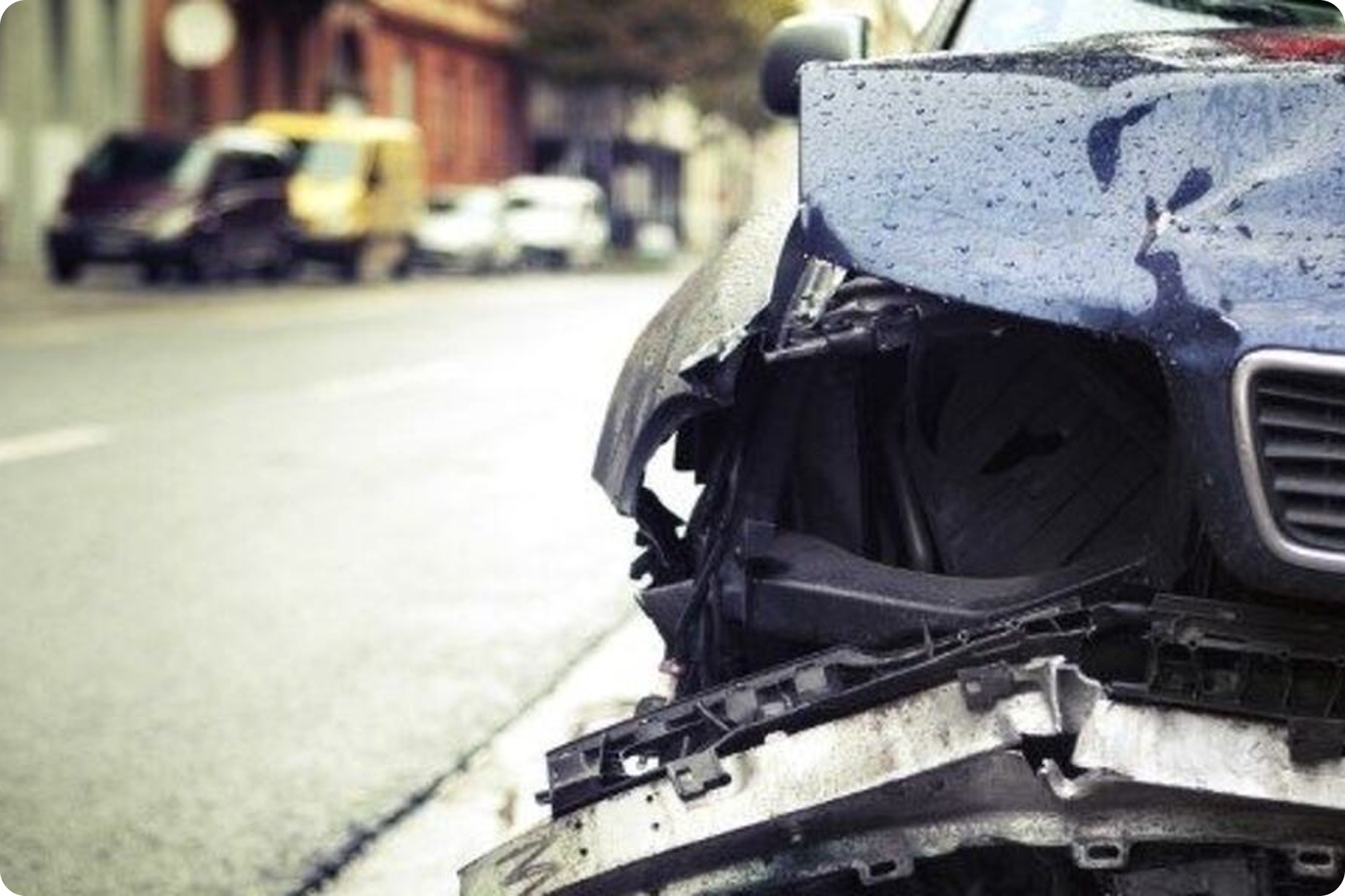 Close-up of a damaged black car, with visible scratches and a broken front bumper, parked on a street with other vehicles in the background.