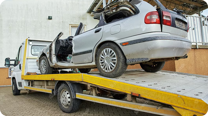 A damaged silver car with an open driver's side door and a broken rear starre and trunk, loaded on a yellow flatbed tow truck, parked on a paved surface near a building.