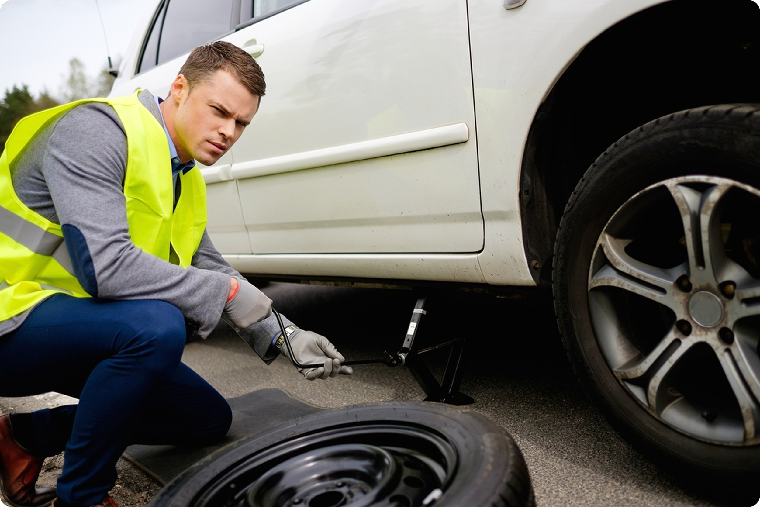 Man wearing a yellow safety vest changing a flat tire on a white car on the side of the road.