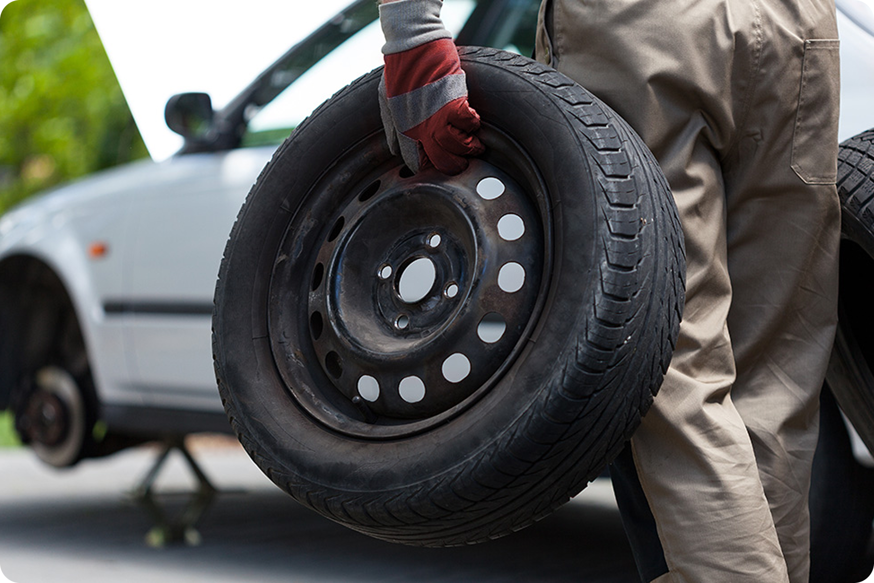 Person wearing beige pants and gray shirt with red gloves holding a car tire outdoors. The car's hood is open in the background.