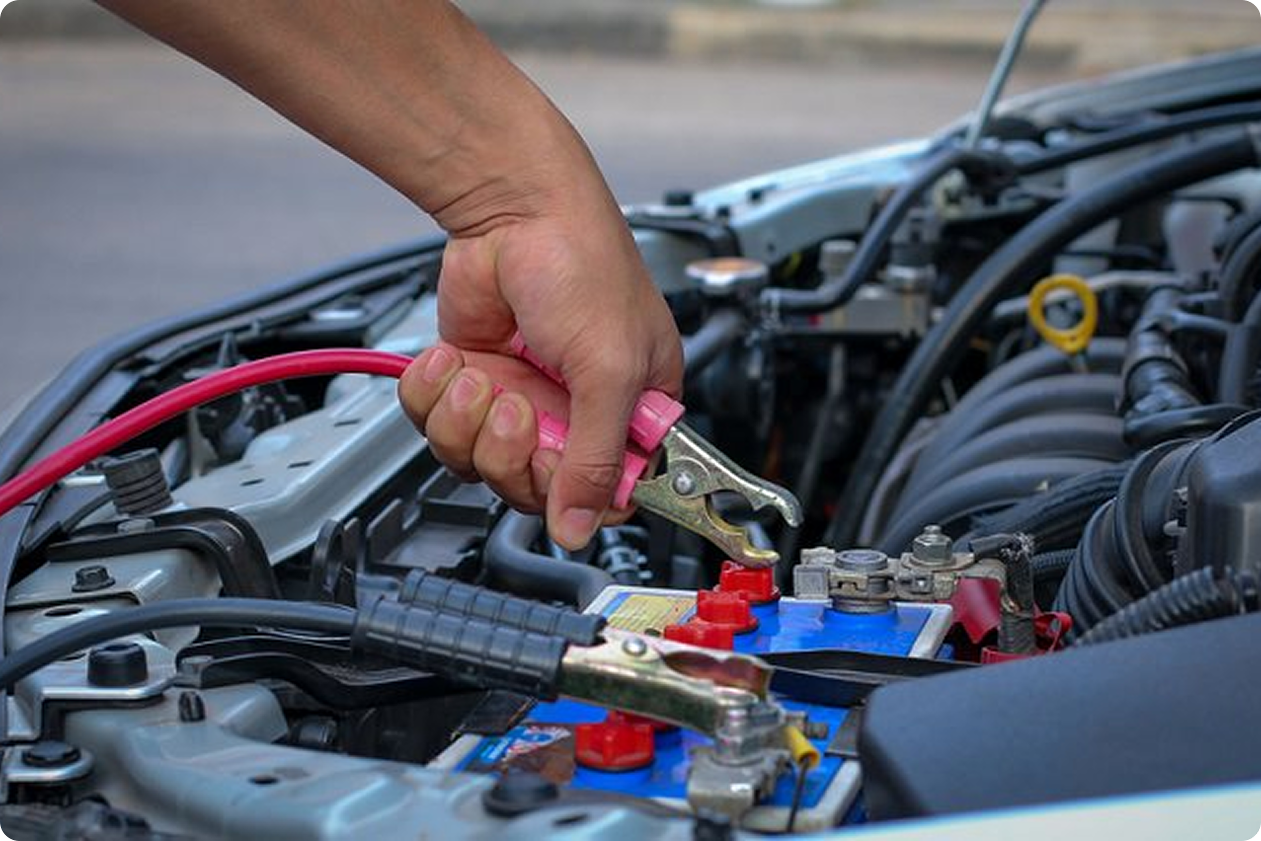 A person using a pink jumper cable to jump start a car. The image shows a close-up of the person's hand connecting the cable to the battery terminal of the car, with the engine of the car, various wires, and components visible.