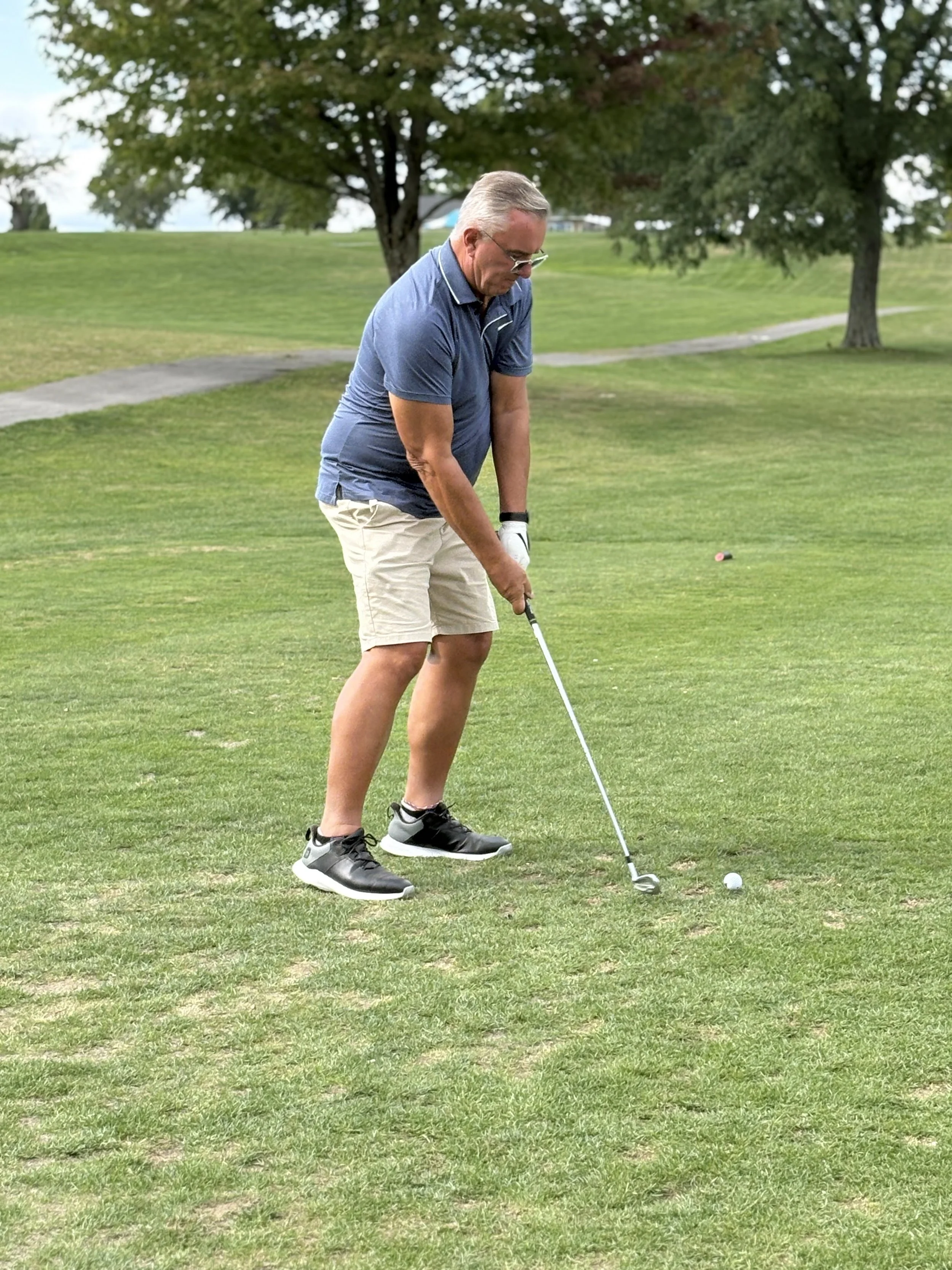 An elderly man in a blue polo shirt, beige shorts, and black shoes playing golf on a green course. He is preparing to hit a golf ball with a club, standing on the grass with trees and a pathway in the background.