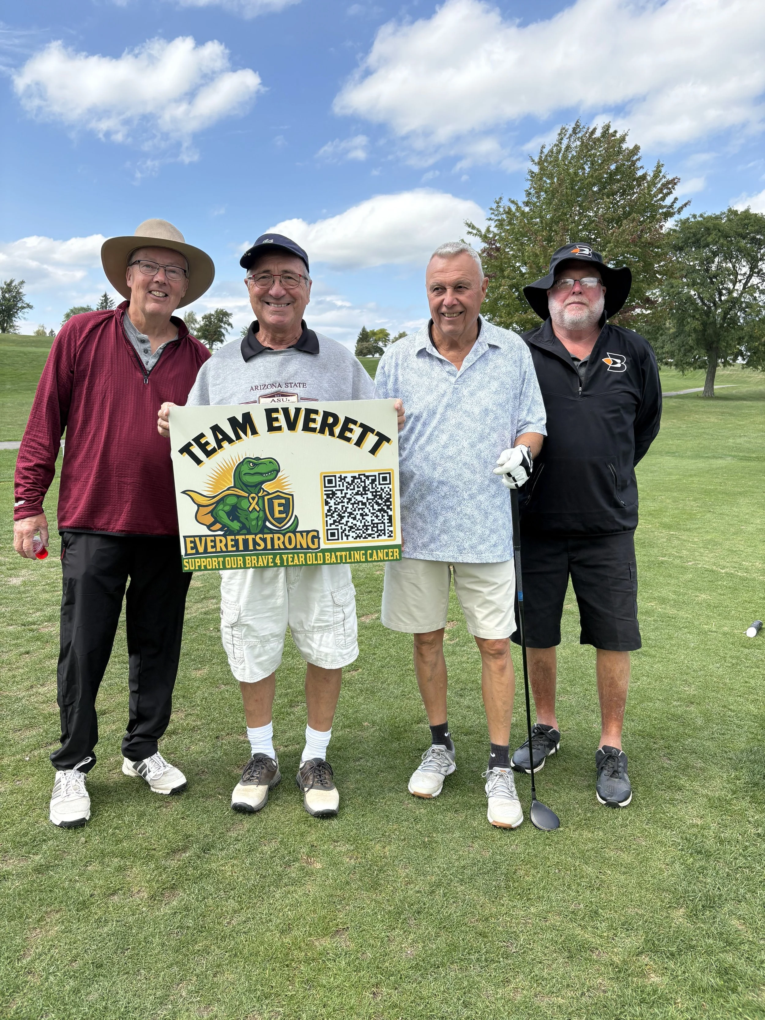 Four older men standing on a golf course, smiling and holding a sign supporting a cancer cause. The men are dressed in casual golf attire with hats and sunglasses. The sign reads 'Team Everett, Everett Strong, Support Our Brave 4 Year Old Battling Ca