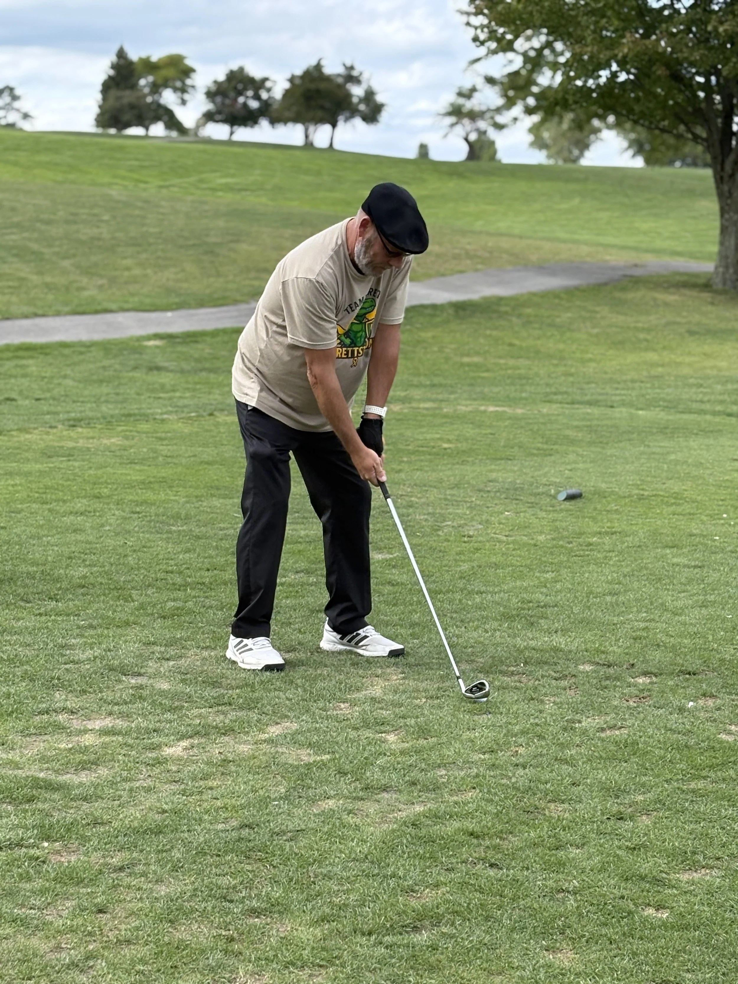 A man playing golf on a grassy course, preparing to hit a golf ball with a club, wearing a beige t-shirt, black pants, and a black beret, with trees and a pathway in the background.