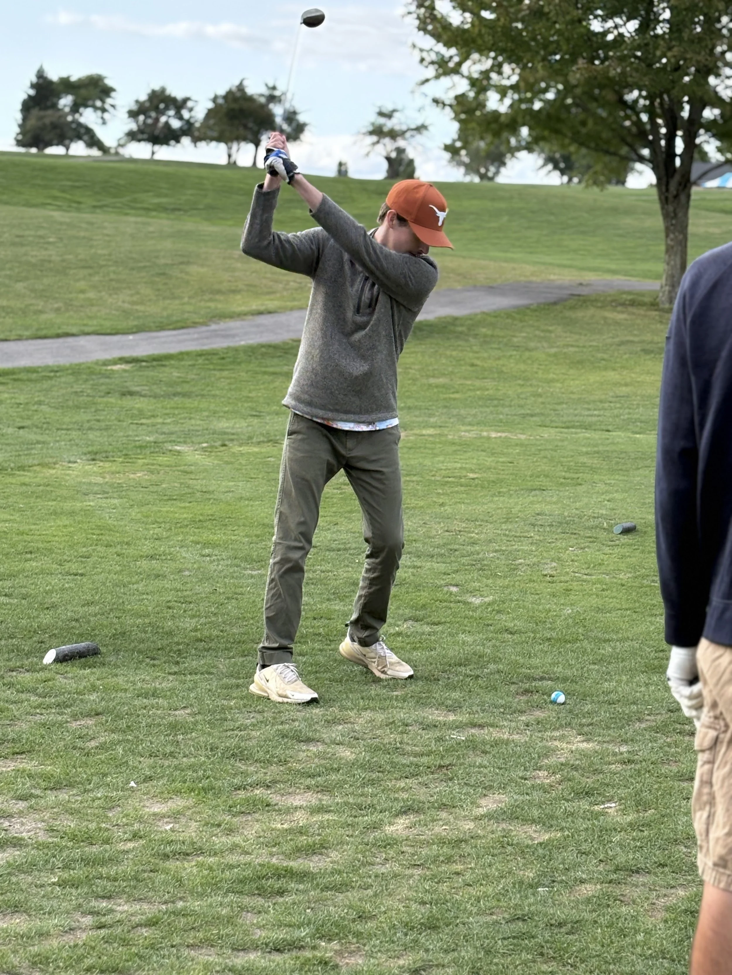 A person in a gray sweater and khaki pants is swinging a golf club at a golf ball on the grass at a golf course. They are wearing a burnt orange cap. Trees and a blue sky are visible in the background.