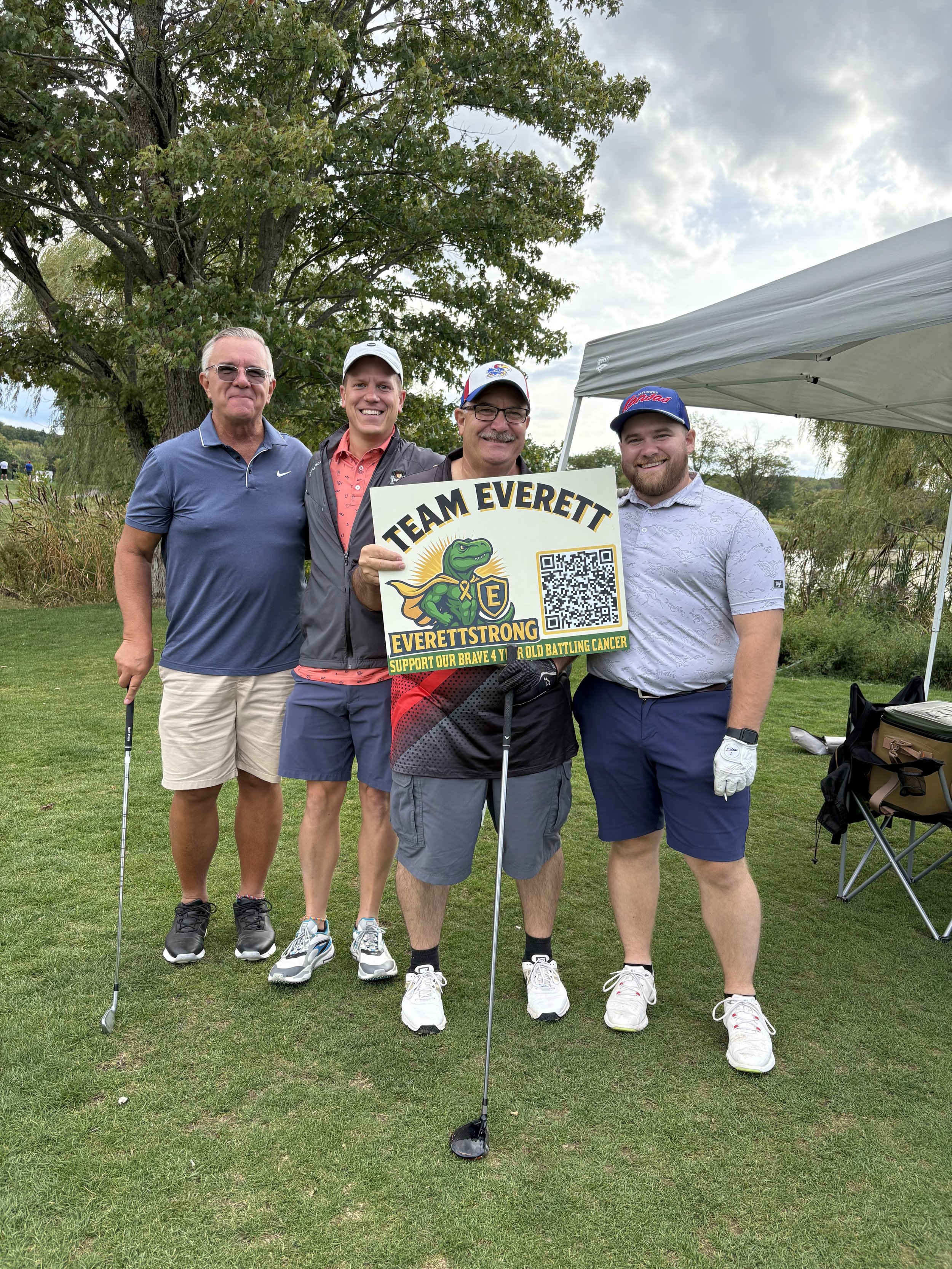 Four men standing together on a golf course, holding a sign that says 'Team Everett, Everett Strong, Support Our Brave 4 Year Old Battling Cancer,' with trees and a cloudy sky in the background.