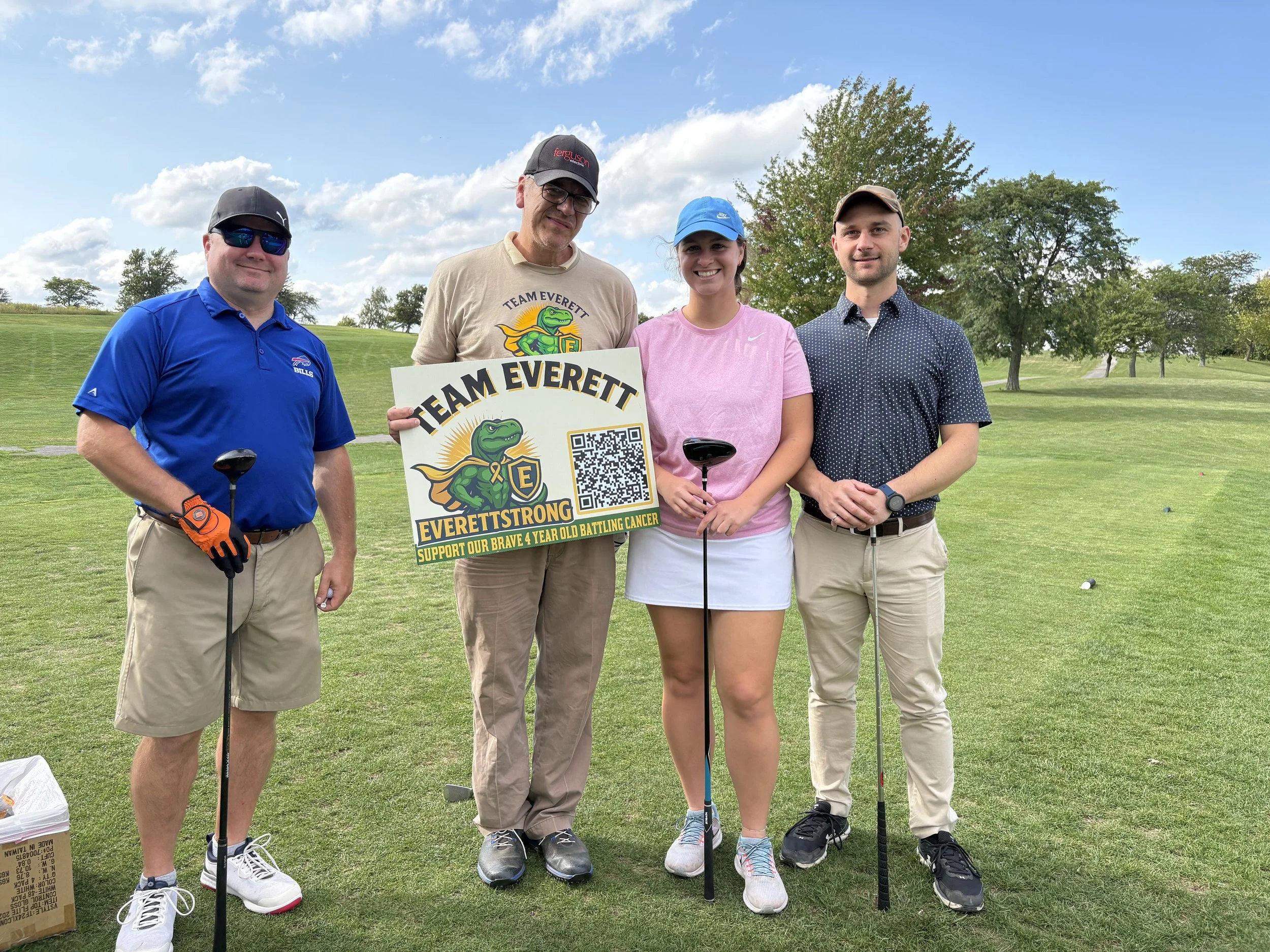 Four people standing on a golf course holding a sign supporting a cancer charity, with trees and a clear blue sky in the background.