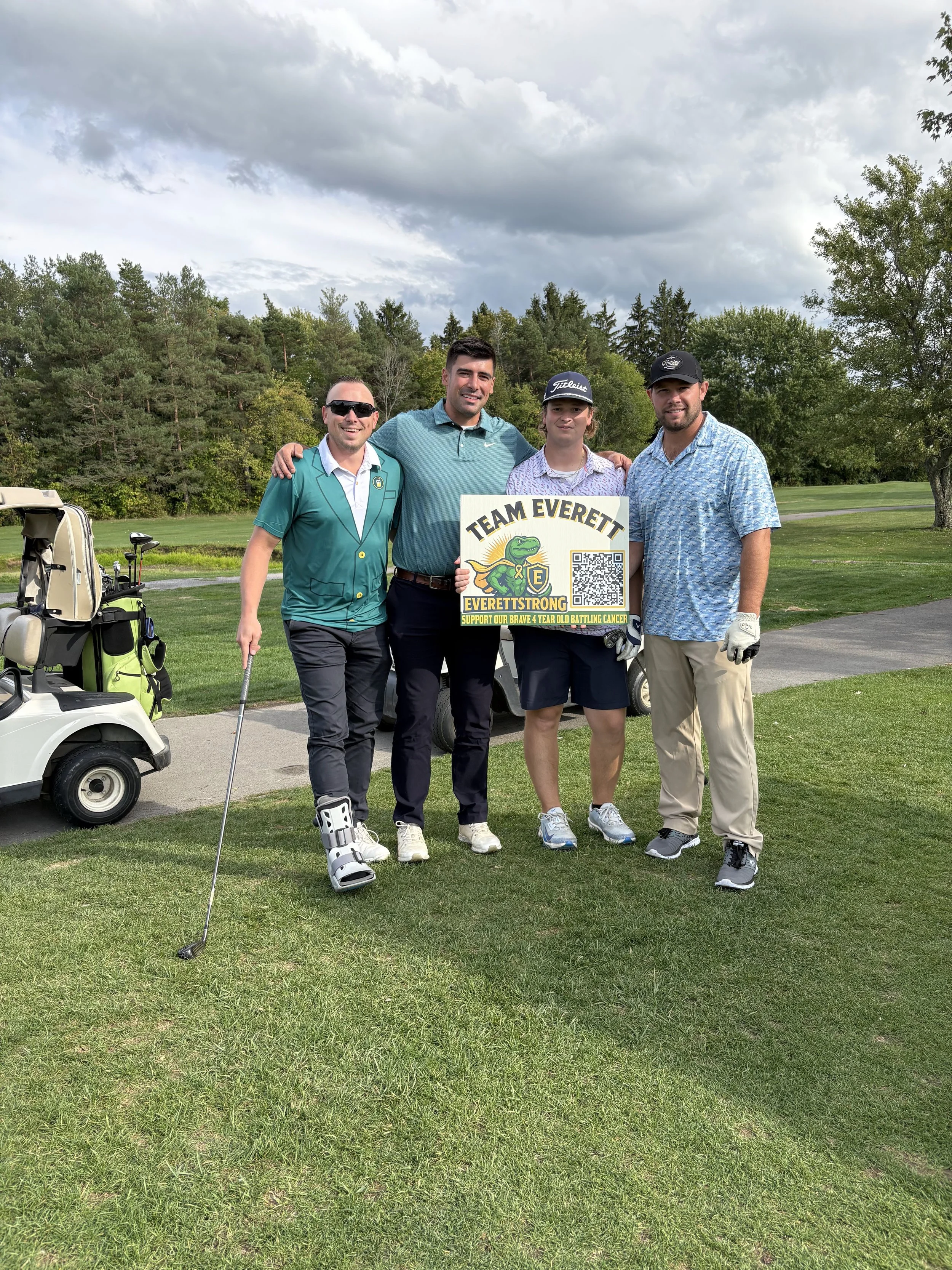 Four men standing on a golf course holding a sign that supports children battling cancer. They are smiling, with golf clubs and a golf cart nearby, and trees and cloudy sky in the background.