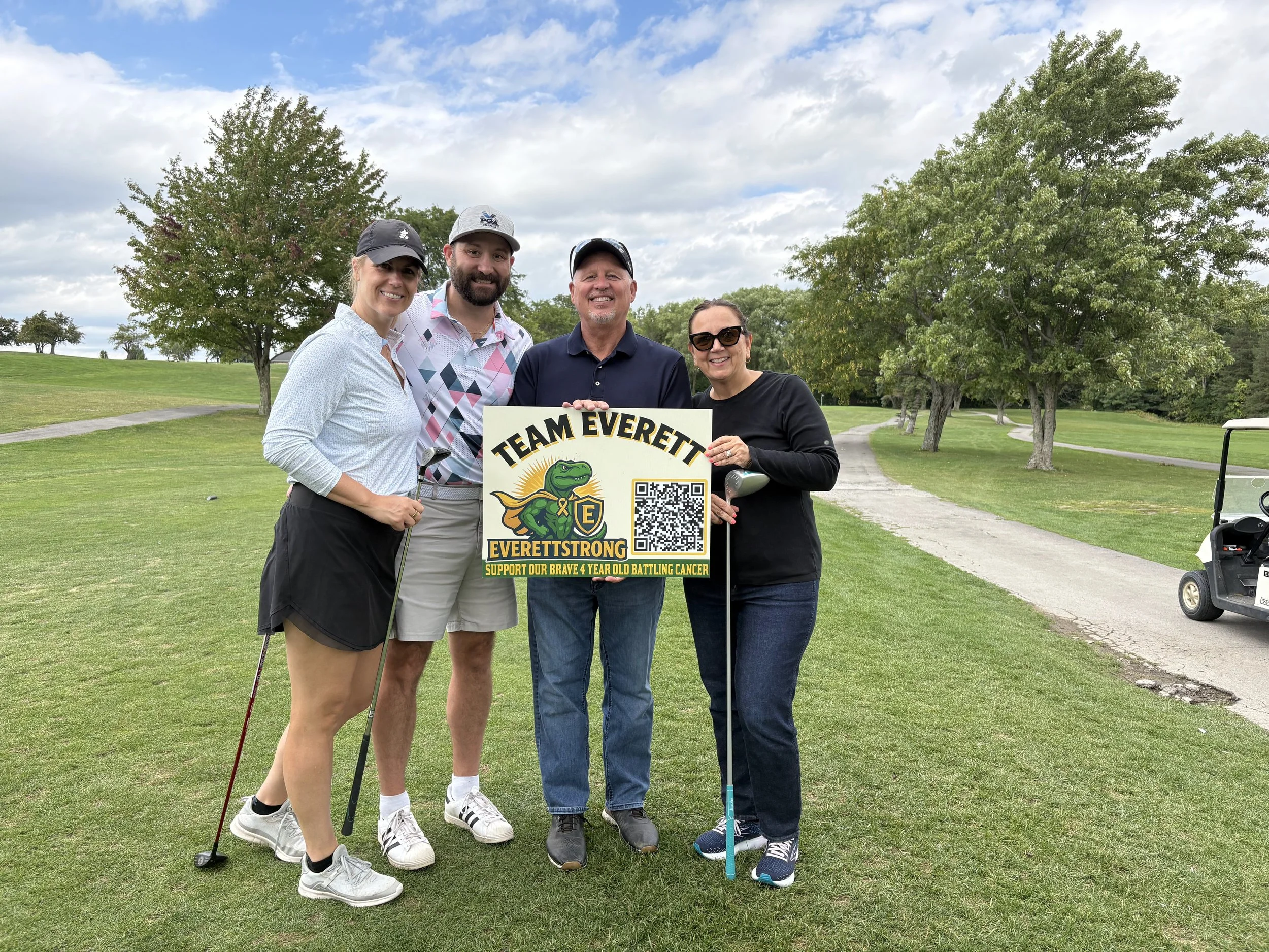 Four people standing on a golf course holding a sign that reads "Team Everett, EverettStrong support our brave 4-year-old battling cancer", with trees and a cloudy sky in the background.