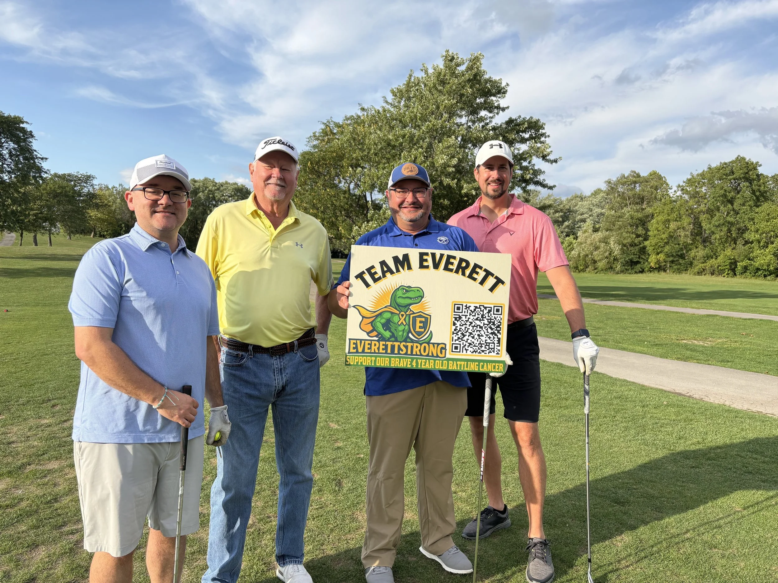 Group of five men standing on a golf course holding a sign that reads 'Team Everett' with a cartoon alligator and QR code, supporting a childhood cancer awareness event.