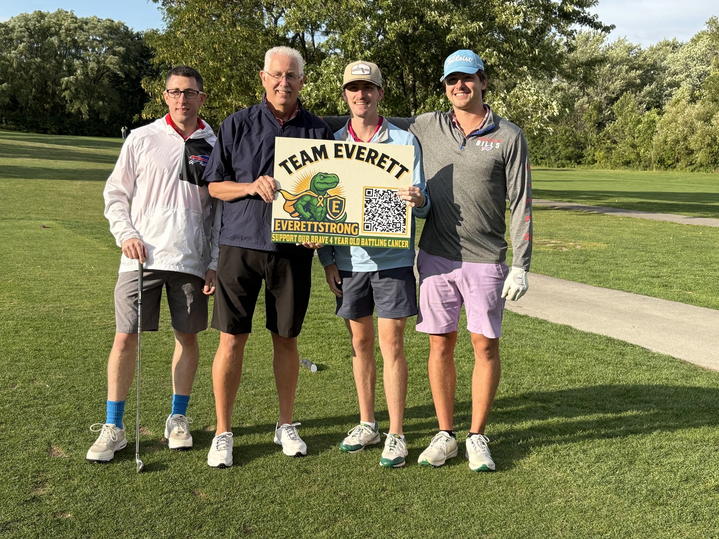 A group of five men standing on a golf course holding a sign that reads "Team Everett" supporting a four-year-old battling cancer, with trees and a partly cloudy sky in the background.