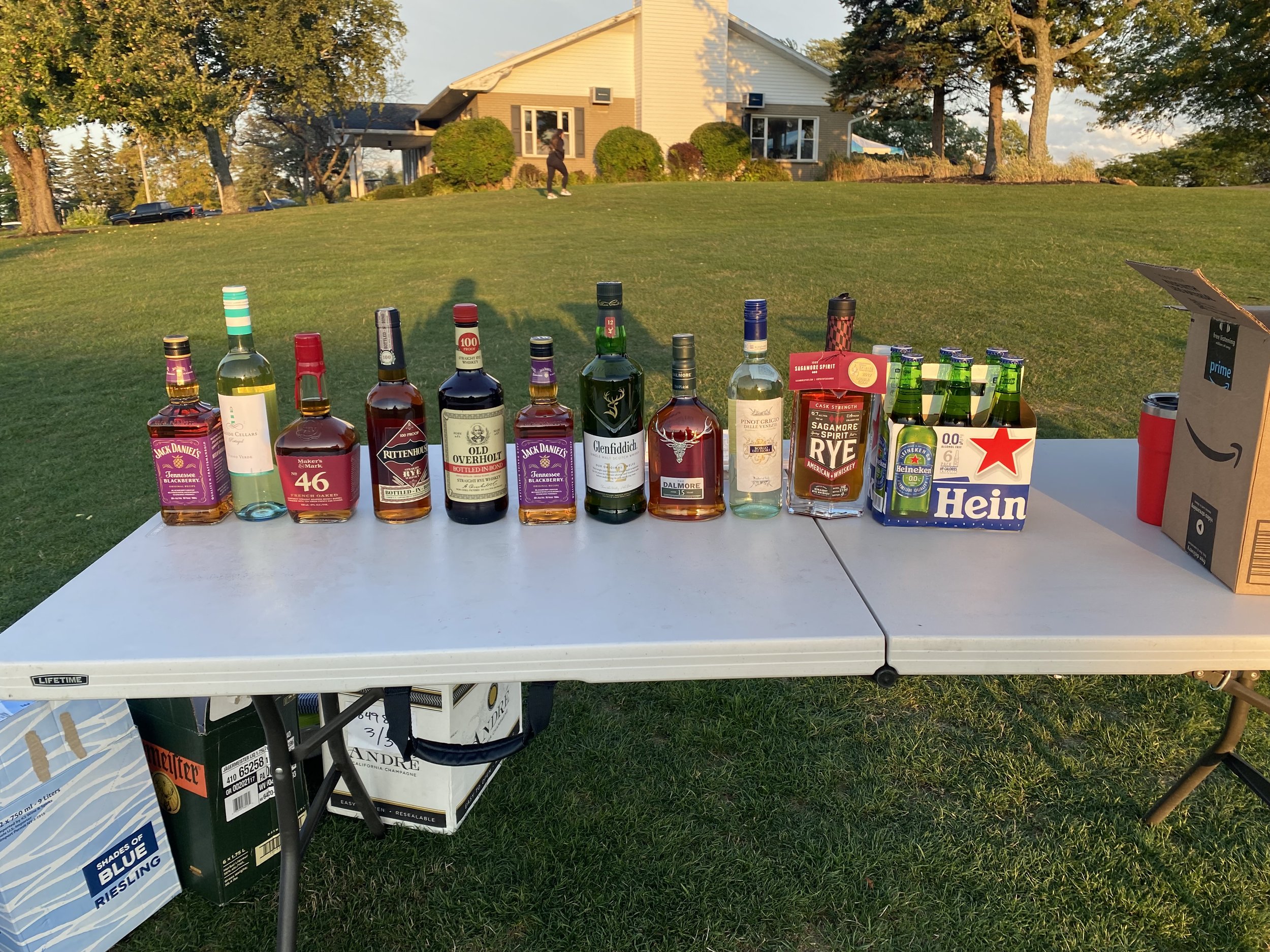 A table with various bottles of alcohol, including whiskey, rum, and flavored liquors, set outdoors on green grass with a house, cars, and trees in the background.