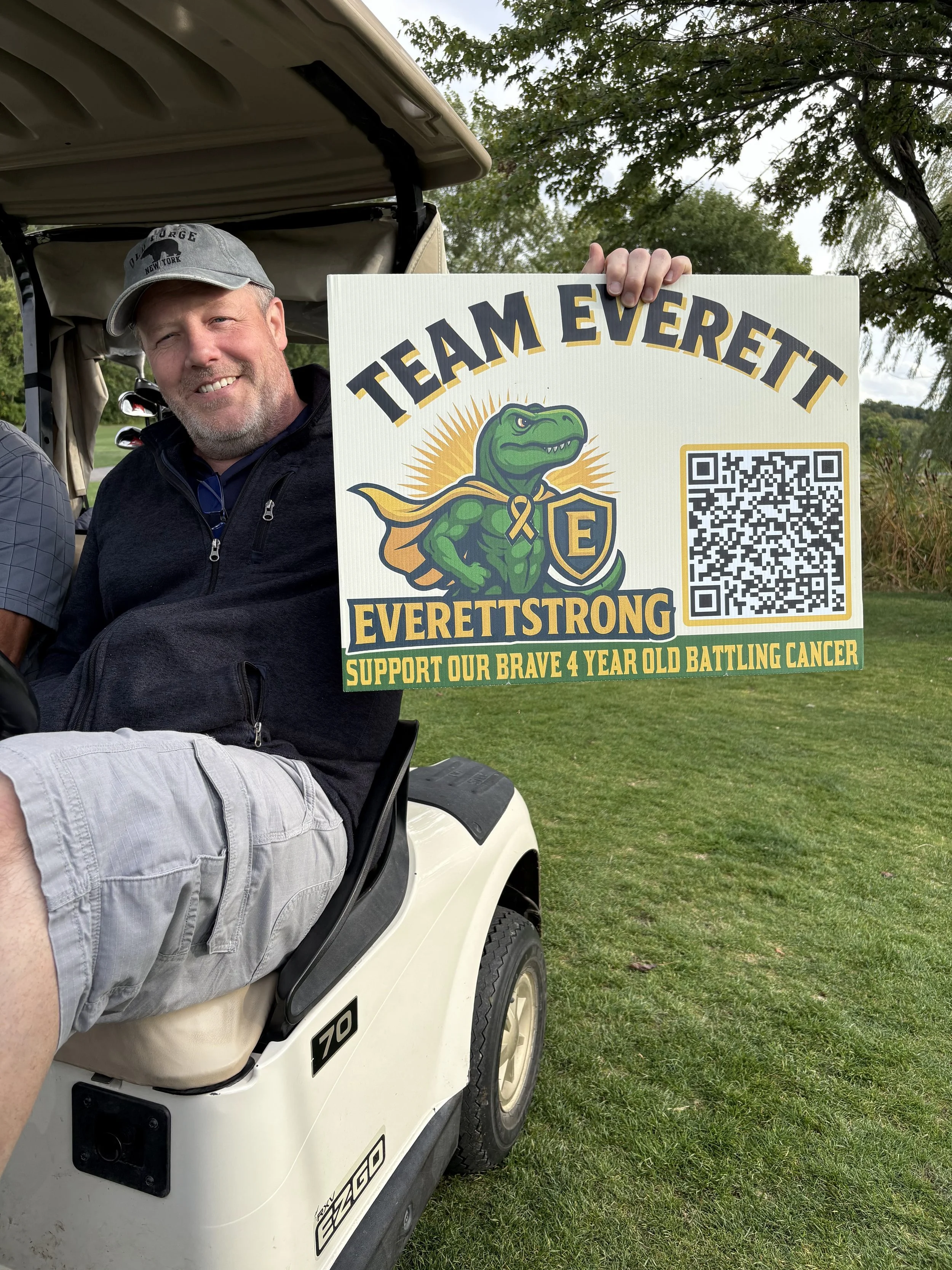 A man with a beard and a gray cap smiling while sitting in a golf cart, holding a sign supporting a childhood cancer fight. The sign reads 'TEAM EVEREST' with a cartoon dinosaur logo, a QR code, and additional text in green and yellow.
