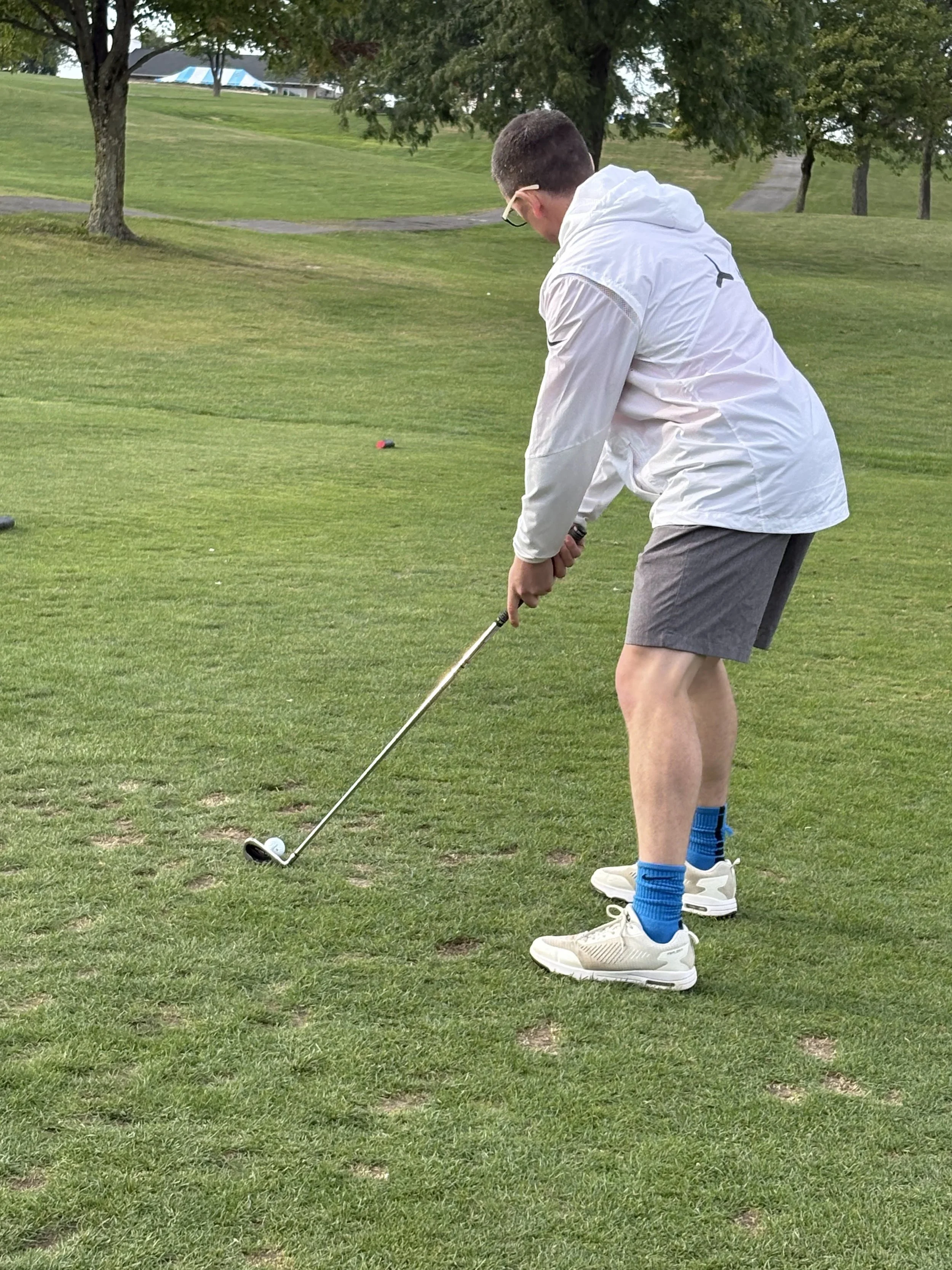 A young man wearing a white jacket, gray shorts, blue socks, and white sneakers is preparing to hit a golf ball on a grassy golf course. He is holding a golf club and bent over slightly, focusing on the ball.