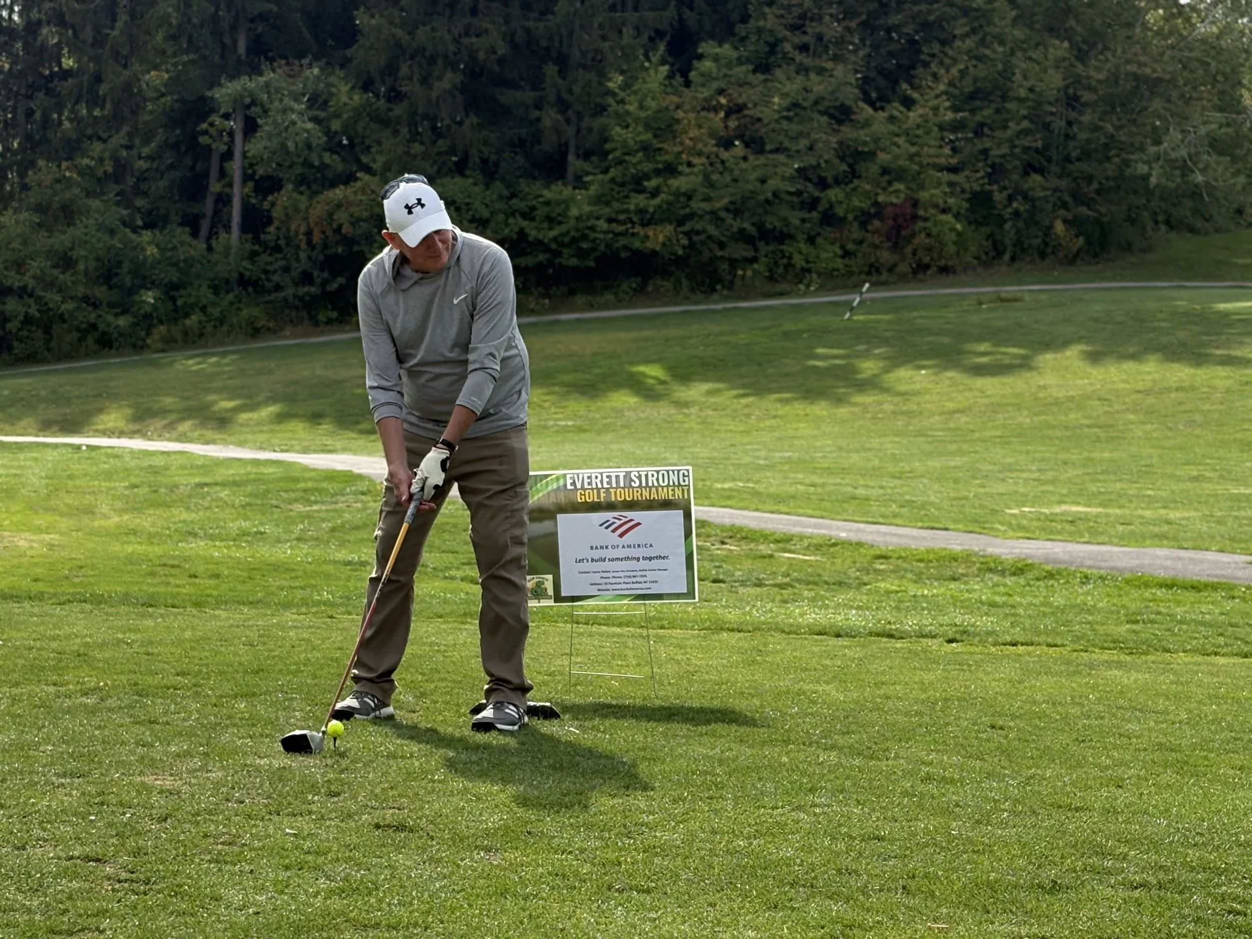 A man in a gray Nike jacket, khaki pants, and a white Under Armour cap is standing on a golf course, preparing to hit a golf ball with a driver club. There is a sign that says 'Everett Strong Golf Tournament' with a Bank of America logo beside him, a