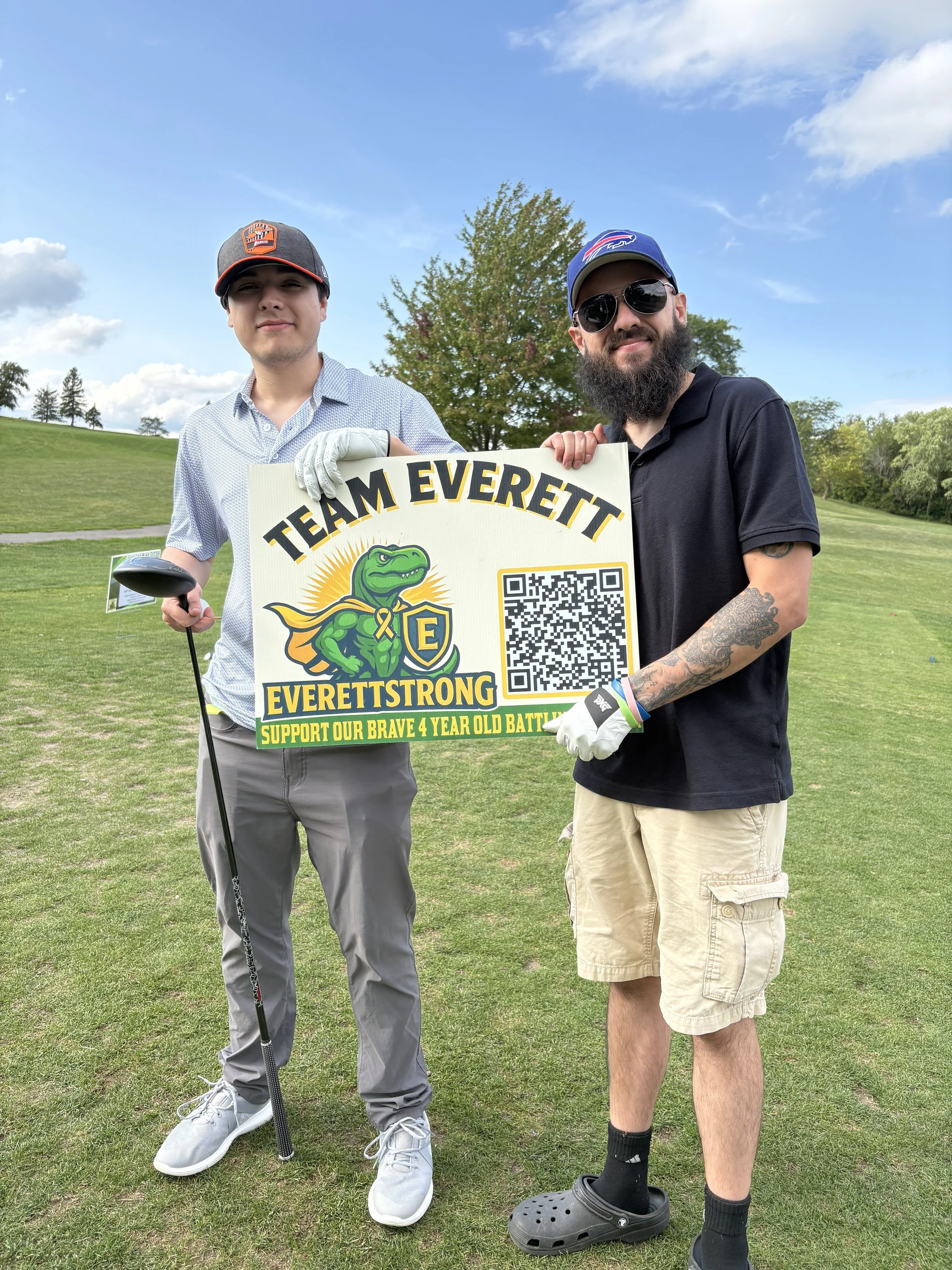 Two young men standing on a golf course, holding a sign that says 'Team Everett', with a cartoon dinosaur and a QR code. One is wearing a baseball cap, light-colored shirt, and holding a golf club. The other has a beard, sunglasses, baseball cap, bla