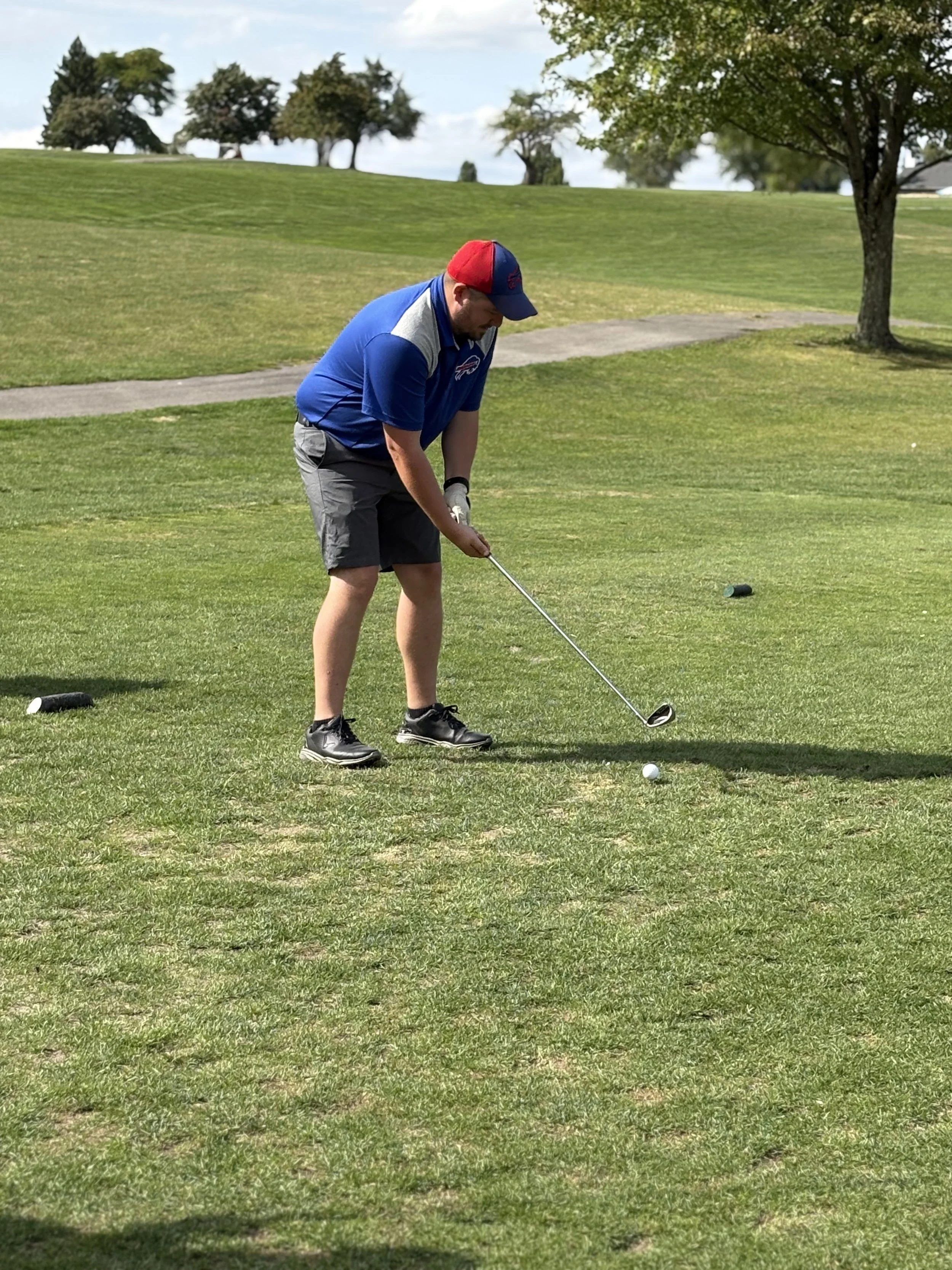A man in a blue and gray golf shirt, gray shorts, and black golf shoes is preparing to hit a golf ball on a golf course. He is wearing a red and blue cap, and there is a golf club in his hands. The golf course is green with trees in the background an