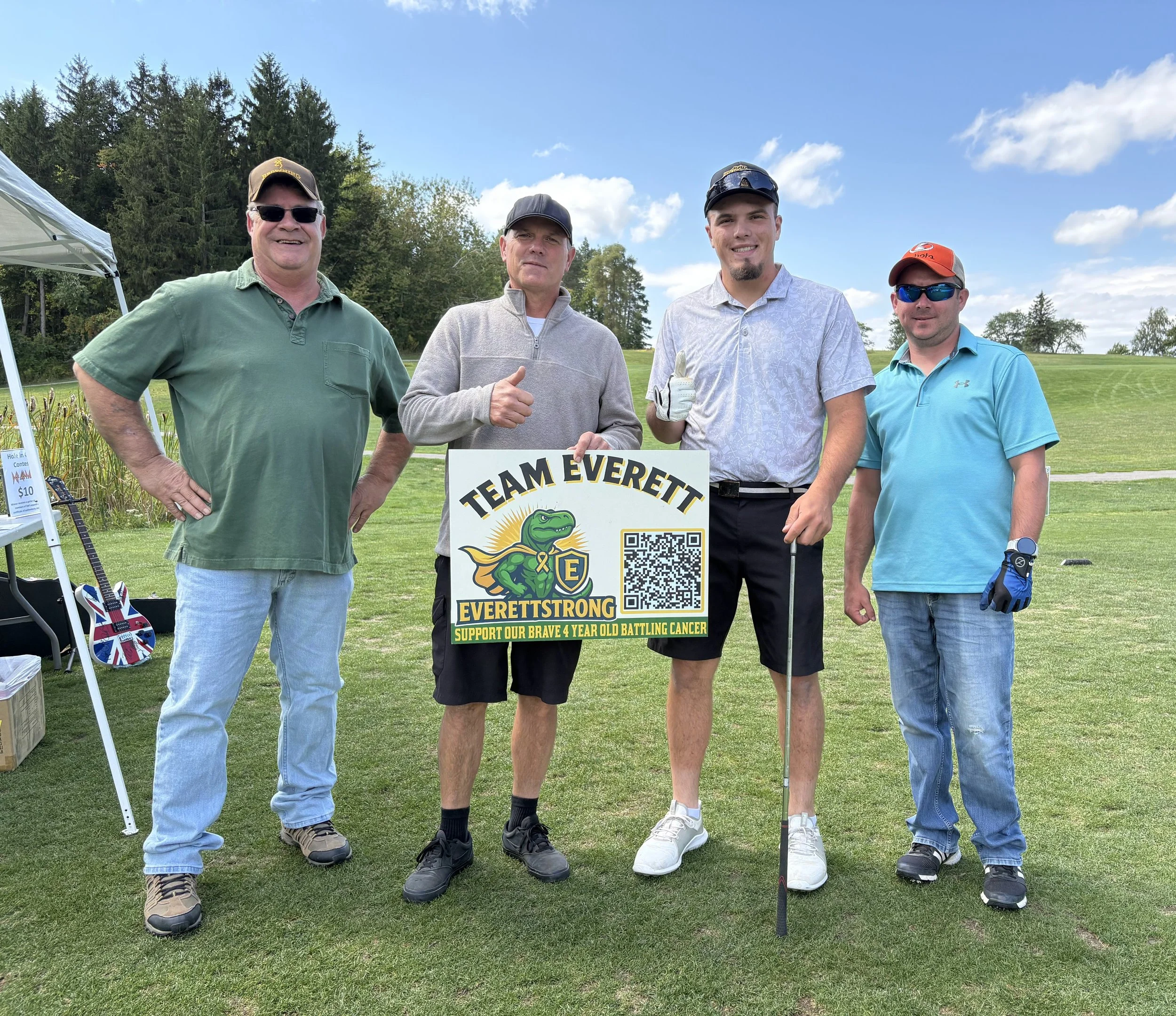 Four men standing outdoors on a golf course, with trees and a blue sky in the background. They are holding a sign that reads 'TEAM EVERETT' with a cartoon alligator and a QR code, supporting a four-year-old battling cancer. The men are wearing casual