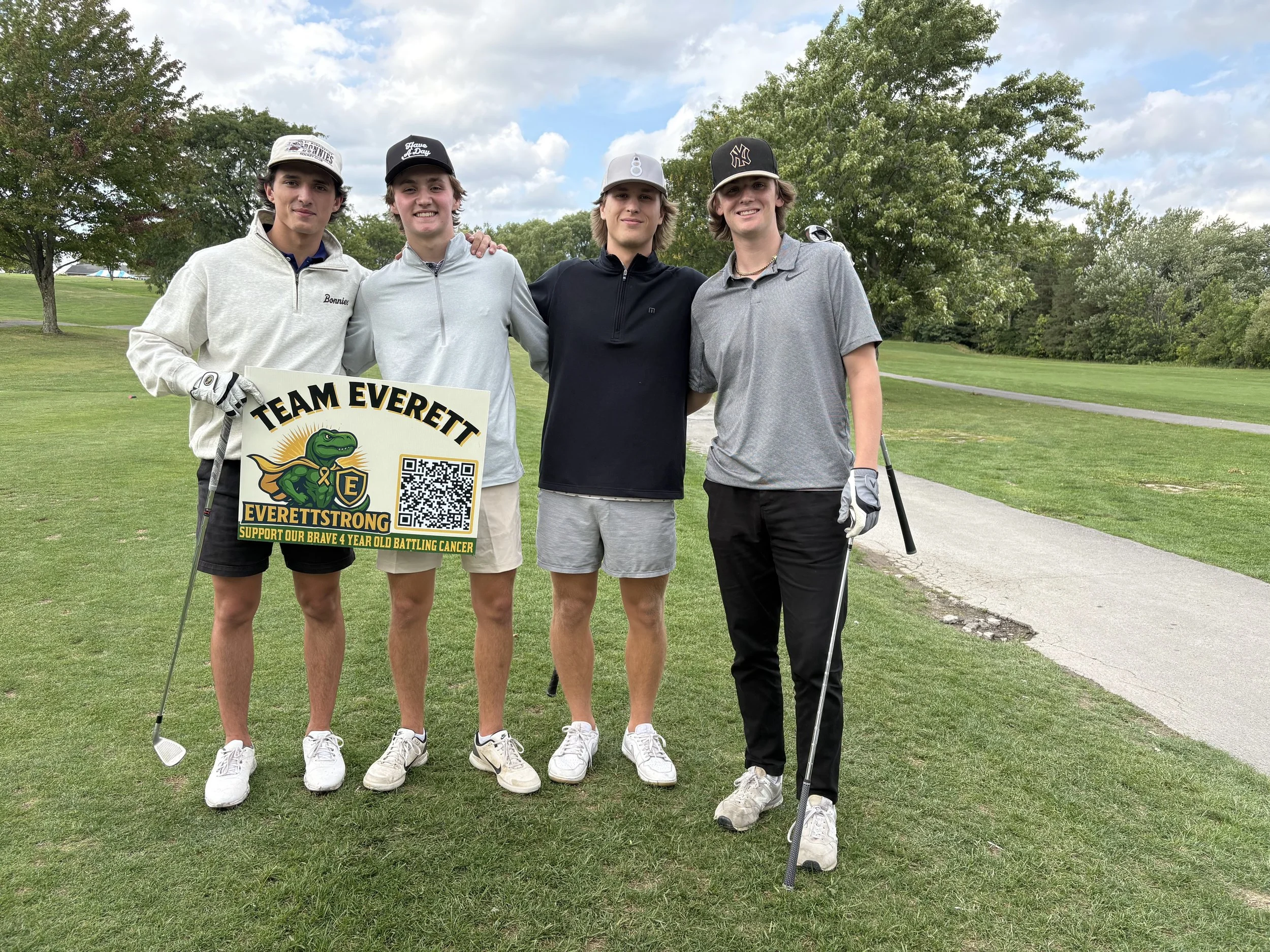 Four young men standing closely together on a golf course, smiling. One is holding a sign with the text "Team Everett" and a cartoon alligator logo, supporting a four-year-old battling cancer. The men are dressed in golf attire, with trees and a blue
