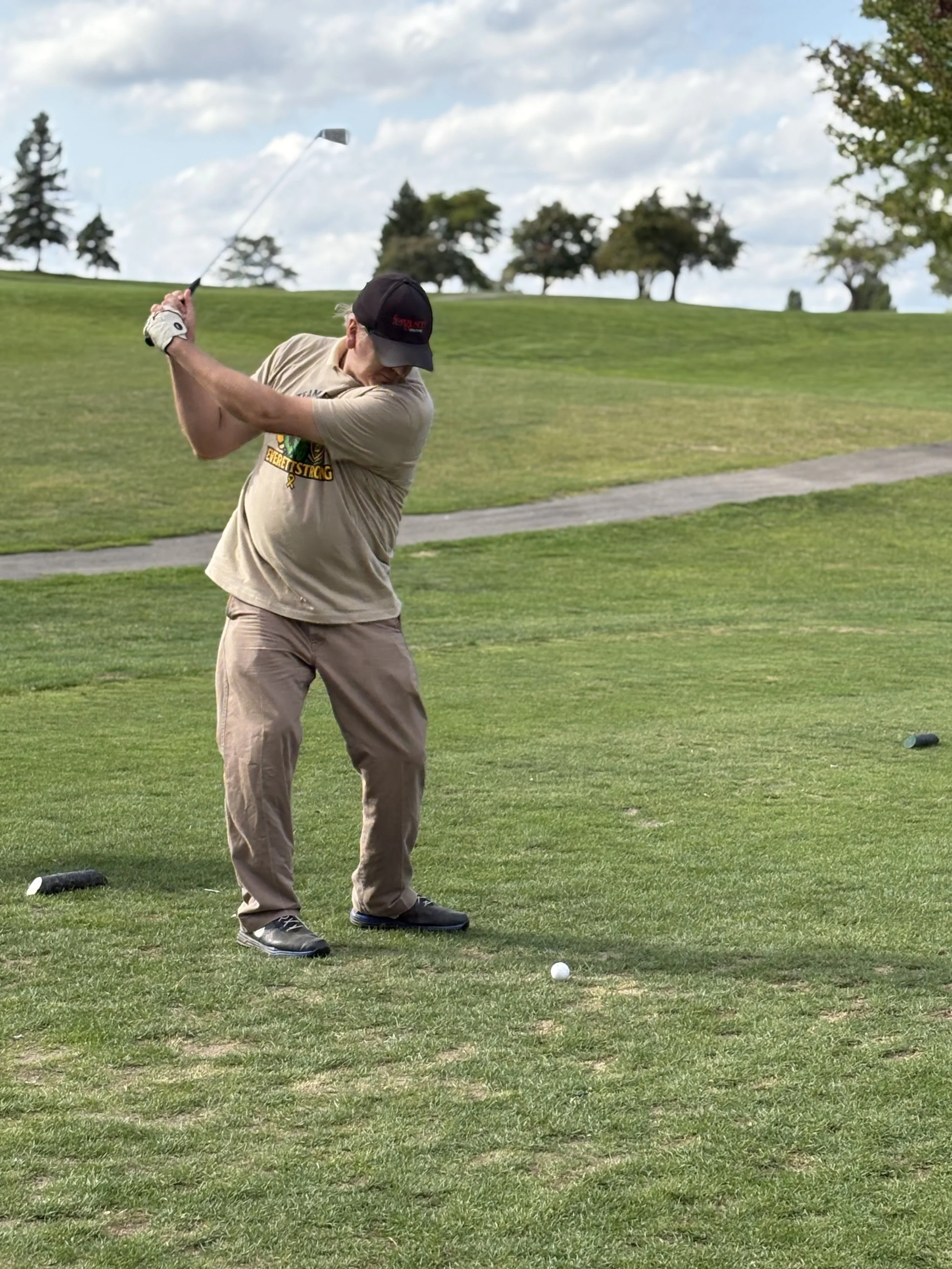 A man wearing a black cap, beige t-shirt, and beige pants swings a golf club at a golf ball on a golf course. Trees and a cloudy sky are visible in the background.
