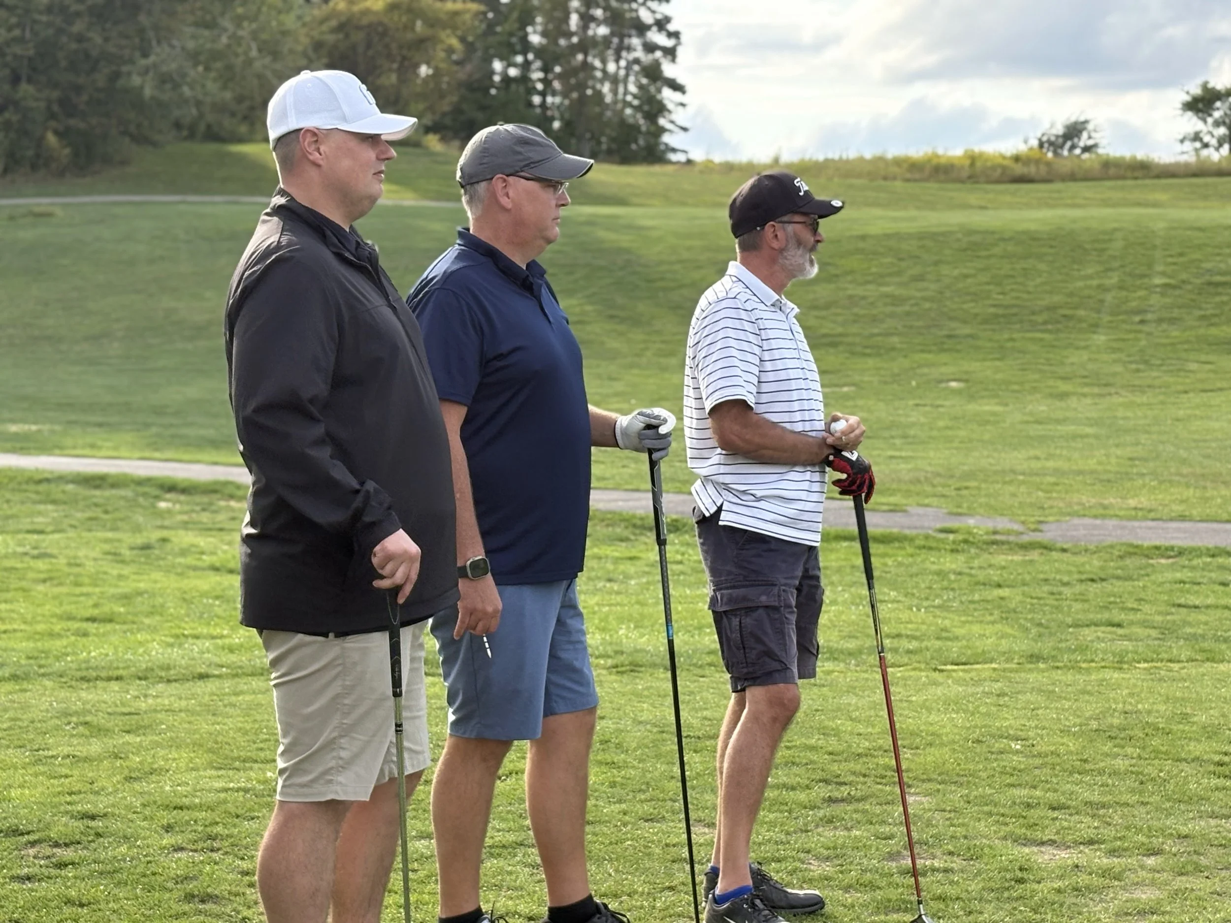 Three middle-aged men standing on a golf course, holding golf clubs, wearing golf hats and casual golf attire, with grass and trees in the background.
