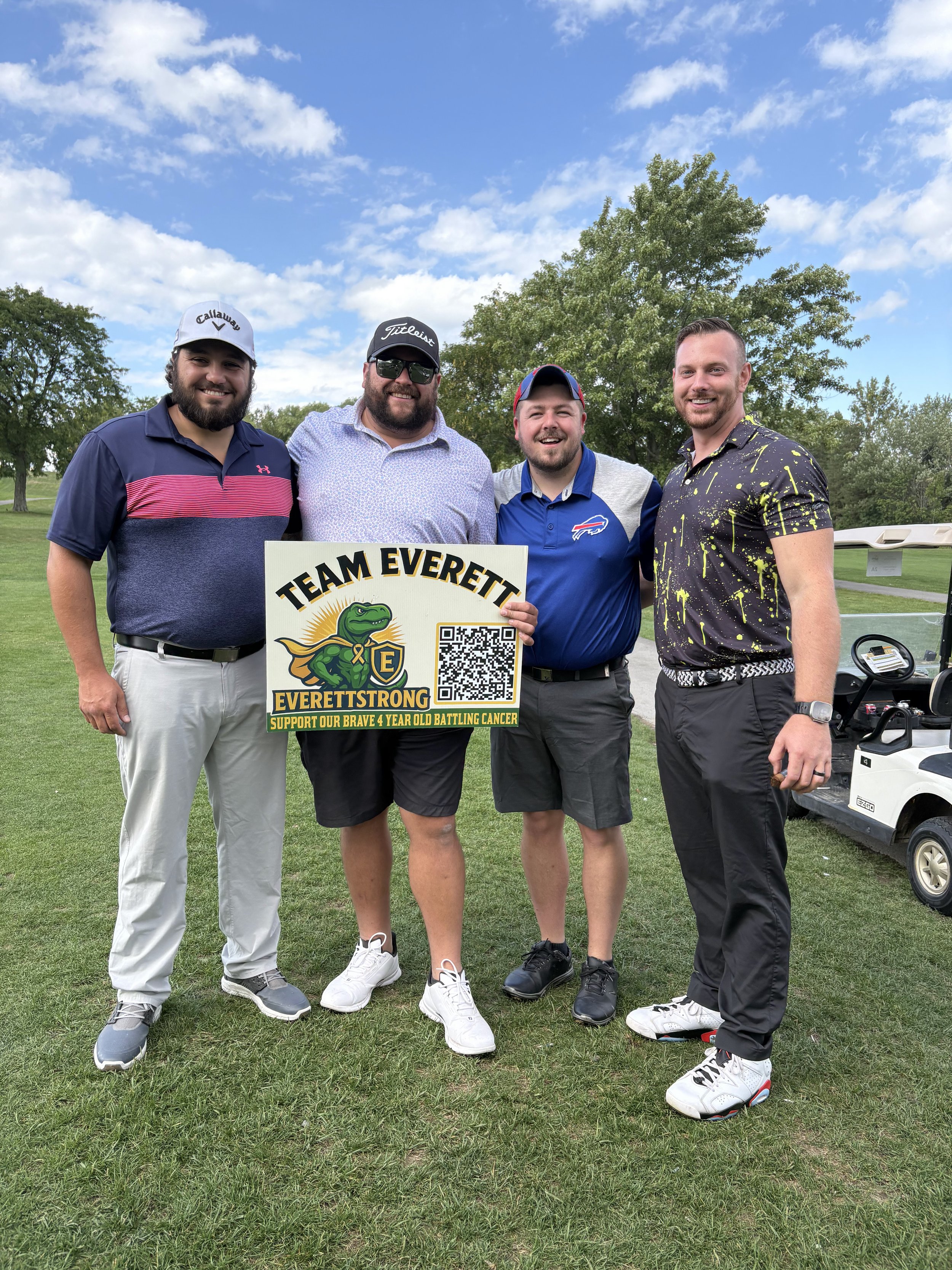 Four men standing on a golf course, holding a sign that reads 'Team Everett, Everett Strong,' supporting a four-year-old battling cancer. The men are smiling and wearing casual golf attire, with a golf cart and trees in the background.