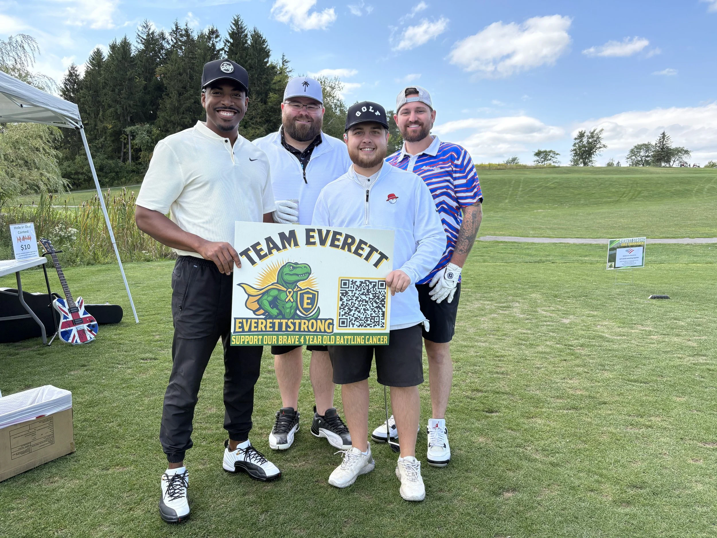 Group of four men standing outdoors on a golf course during daytime, holding a sign supporting a child fighting cancer. They are smiling and dressed in golf attire, with trees and a blue sky in the background.