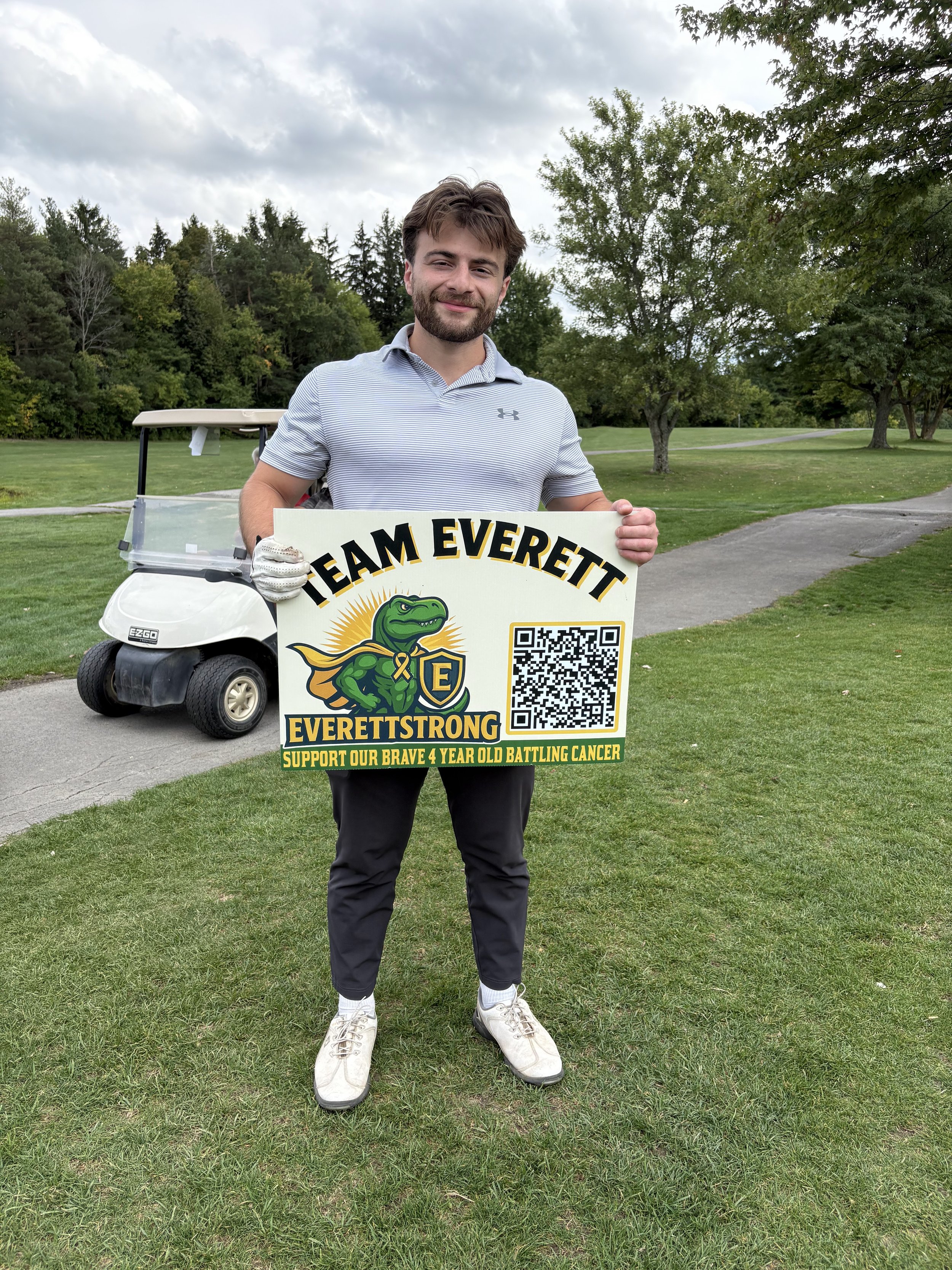 A young man with a beard and brown hair, wearing a striped polo shirt, black pants, and white sneakers, stands on a golf course holding a large sign that reads 'TEAM EVERETT' with a cartoon dinosaur logo and a QR code. A golf cart is visible in the b