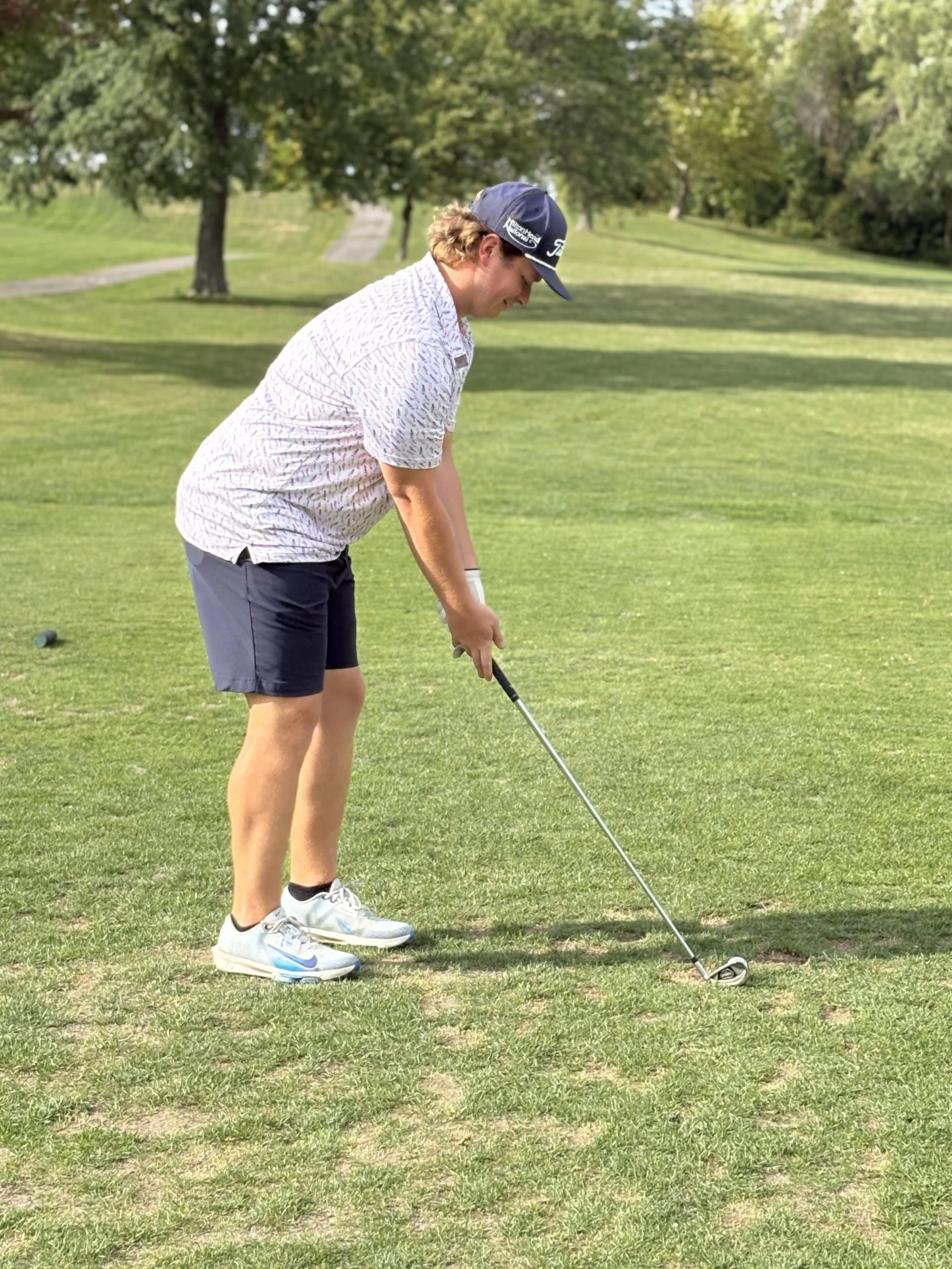 A man in a baseball cap and casual golf attire preparing to hit a golf ball on a grassy course with trees in the background.