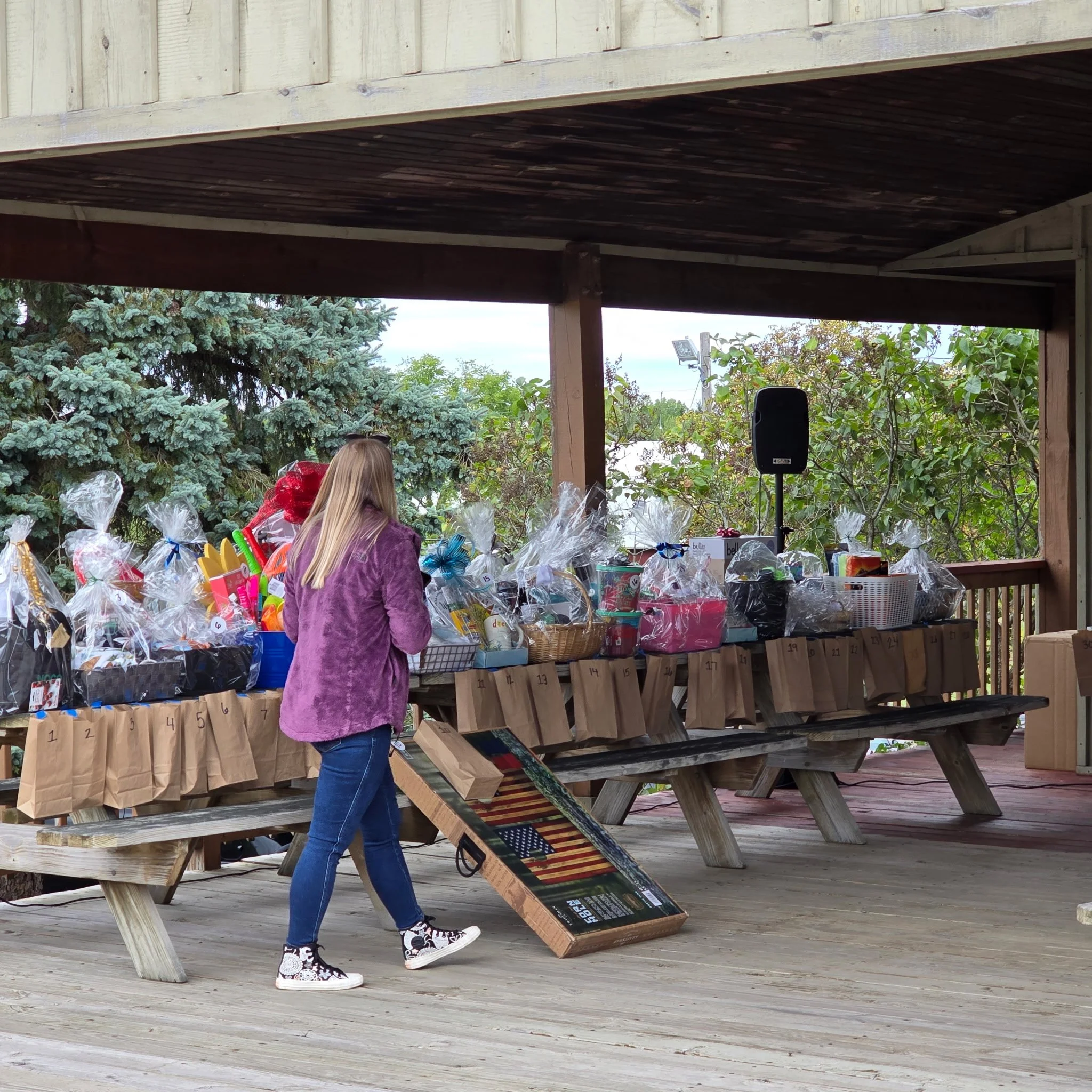A woman with long blonde hair wearing a purple jacket, black and white sneakers, and blue jeans, is shopping for gifts at an outdoor market or store during daytime. She is looking at a table filled with wrapped gifts and gift bags. The background fea