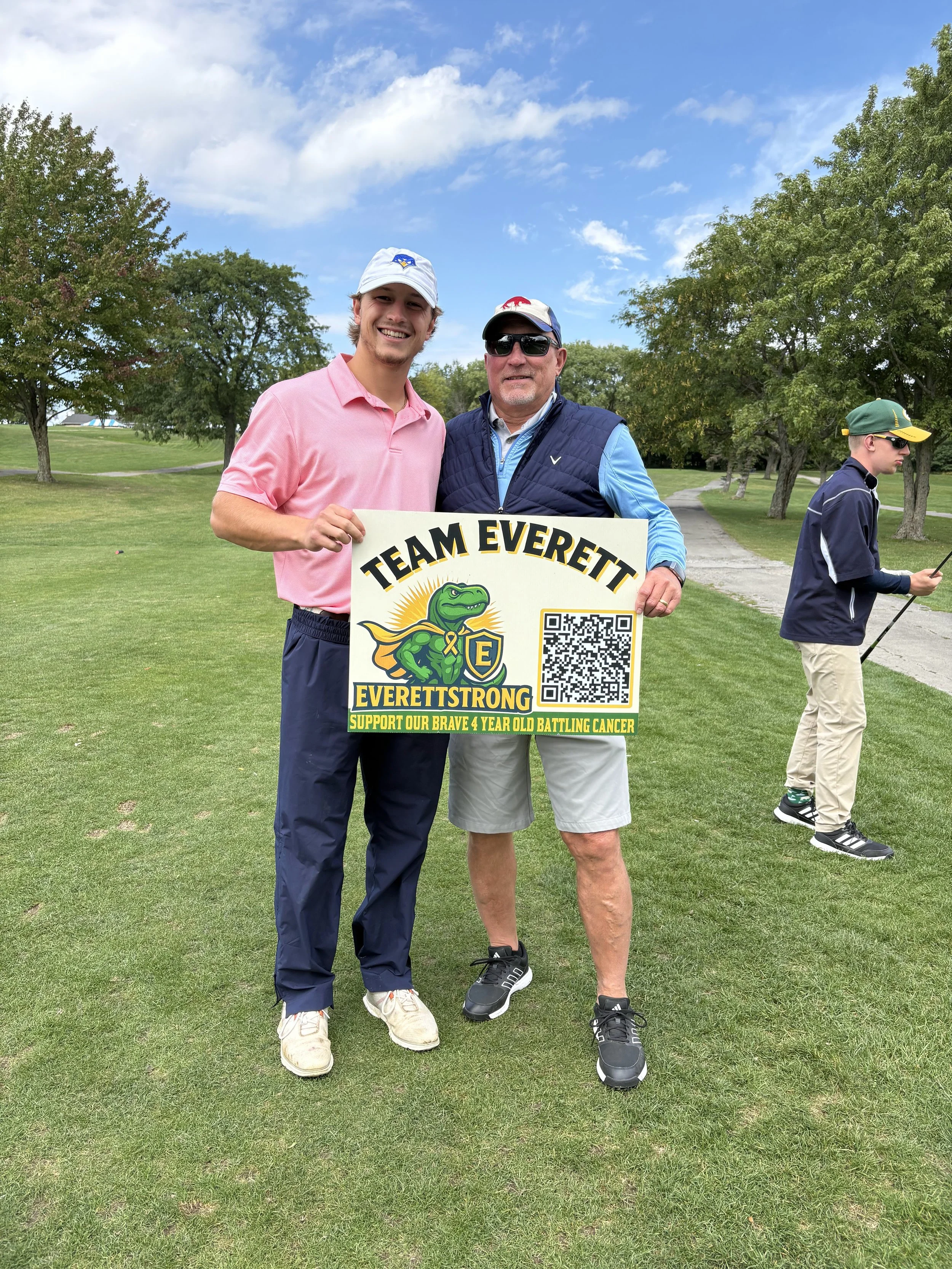 Two men standing outdoors on a grassy area holding a sign that reads 'TEAM EVEREST EVETTSTRONG SUPPORT OUR BRAVE 4 YEAR OLD BATTLING CANCER.' One man is wearing a pink shirt and white cap, while the other is dressed in a blue vest, shorts, and sungla