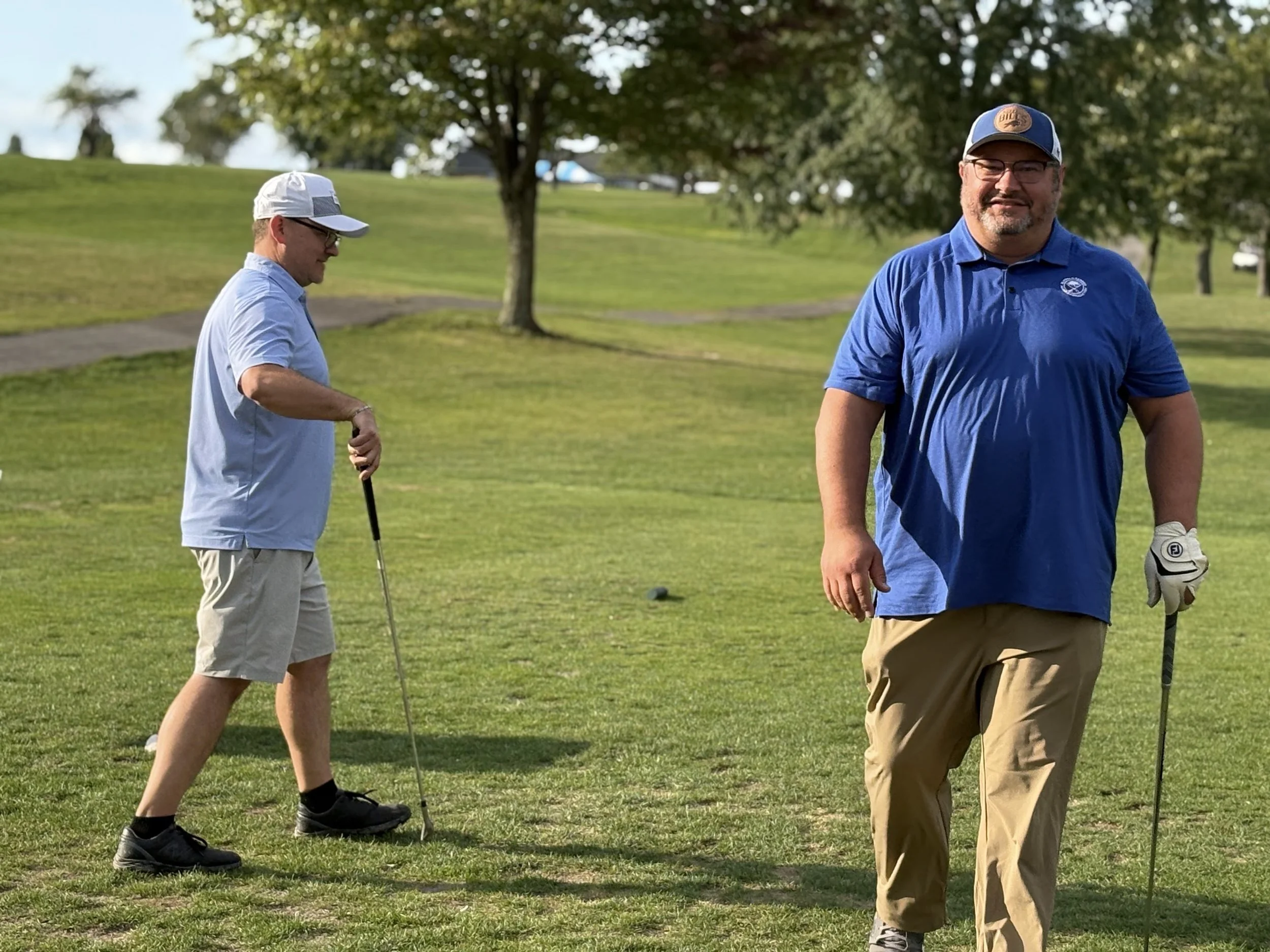 Two men on a golf course, one holding a golf club and the other walking, enjoying a sunny day with green grass and trees in the background.