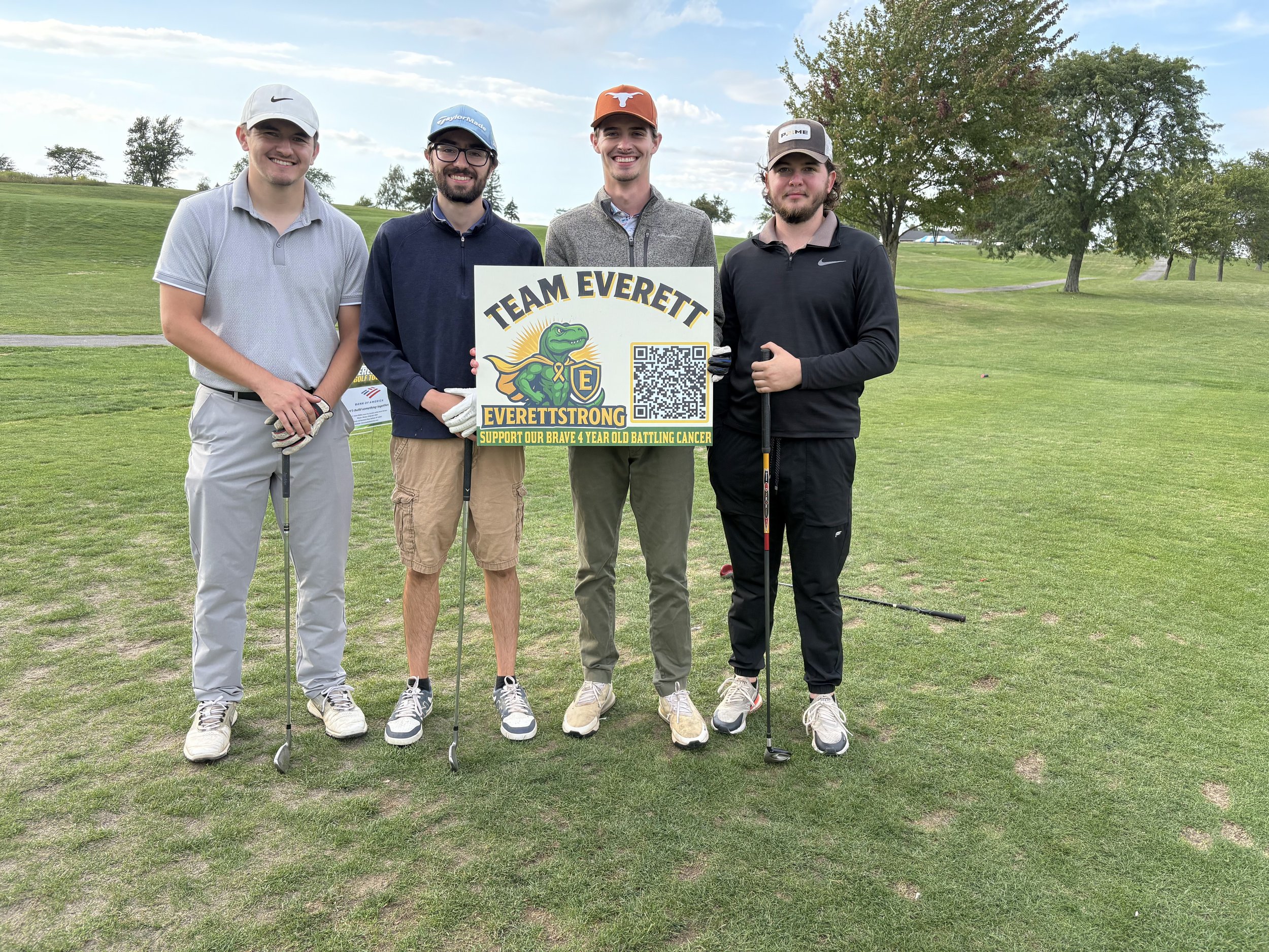 A group of five young men standing on a golf course, holding a sign that reads 'Team Everett, EverettStrong, Support Our Brave 4 Year Old Battling Cancer.' They are dressed in golf attire, wearing hats and holding golf clubs. The background features 