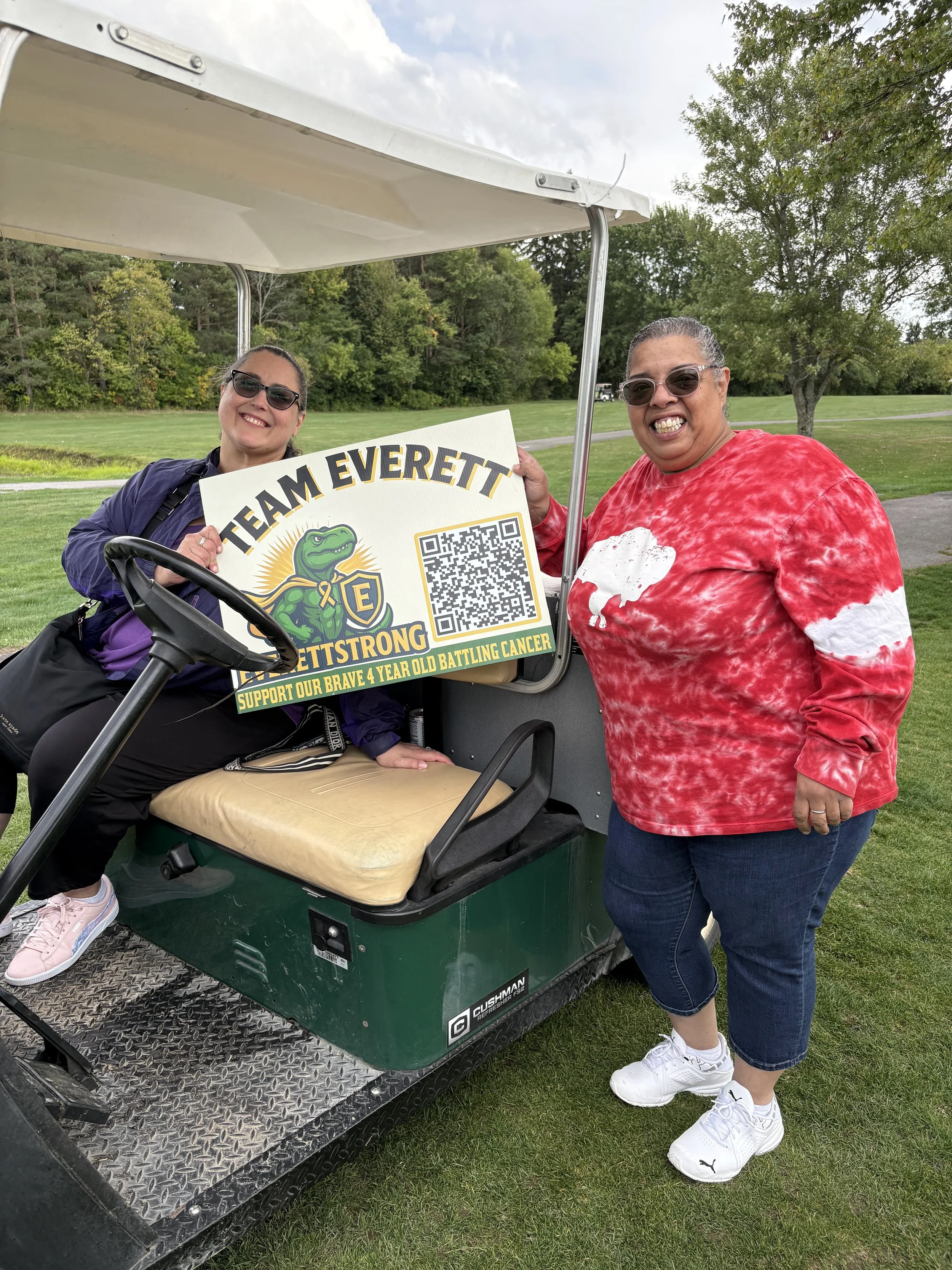 Two women posing next to a golf cart holding a sign supporting breast cancer awareness with a QR code. One is sitting in the golf cart and the other is standing beside it outside on a grassy area with trees in the background.