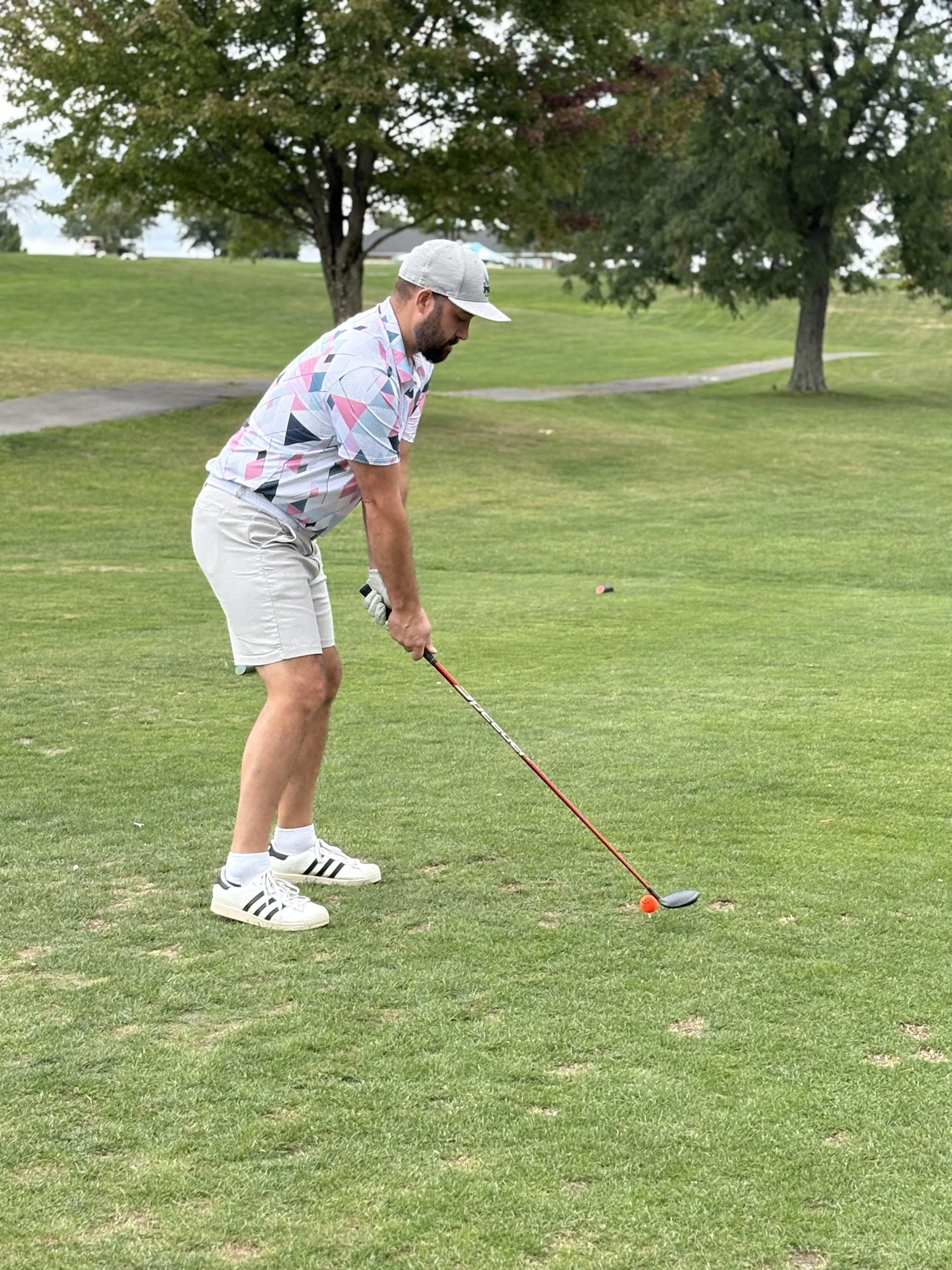 A man in a patterned shirt, shorts, and a gray cap prepares to hit a golf ball on a grassy golf course with trees in the background.