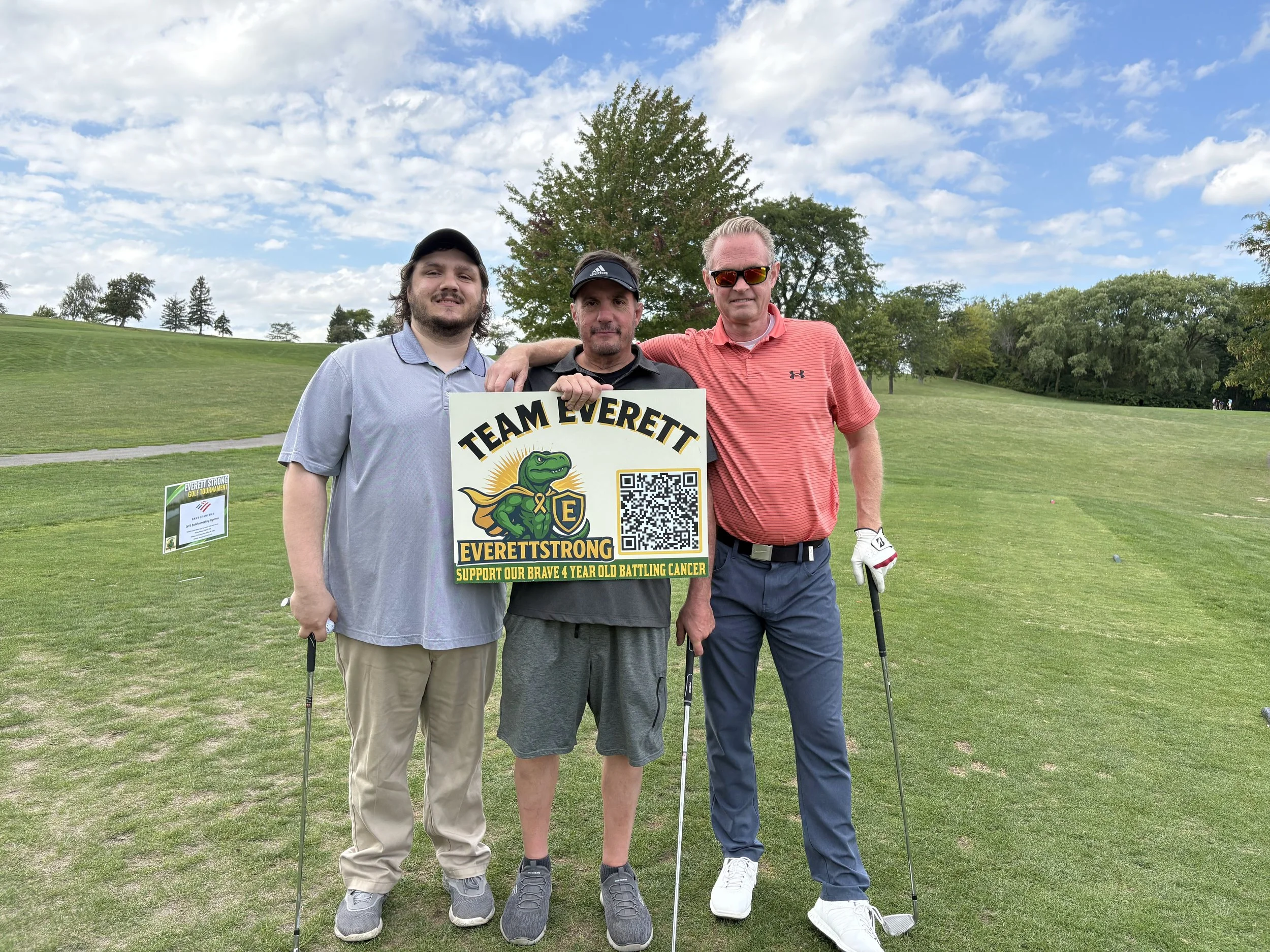 Three men standing on a golf course holding a sign that reads 'Team Everett,' supporting a child battling cancer, with trees and a partly cloudy sky in the background.