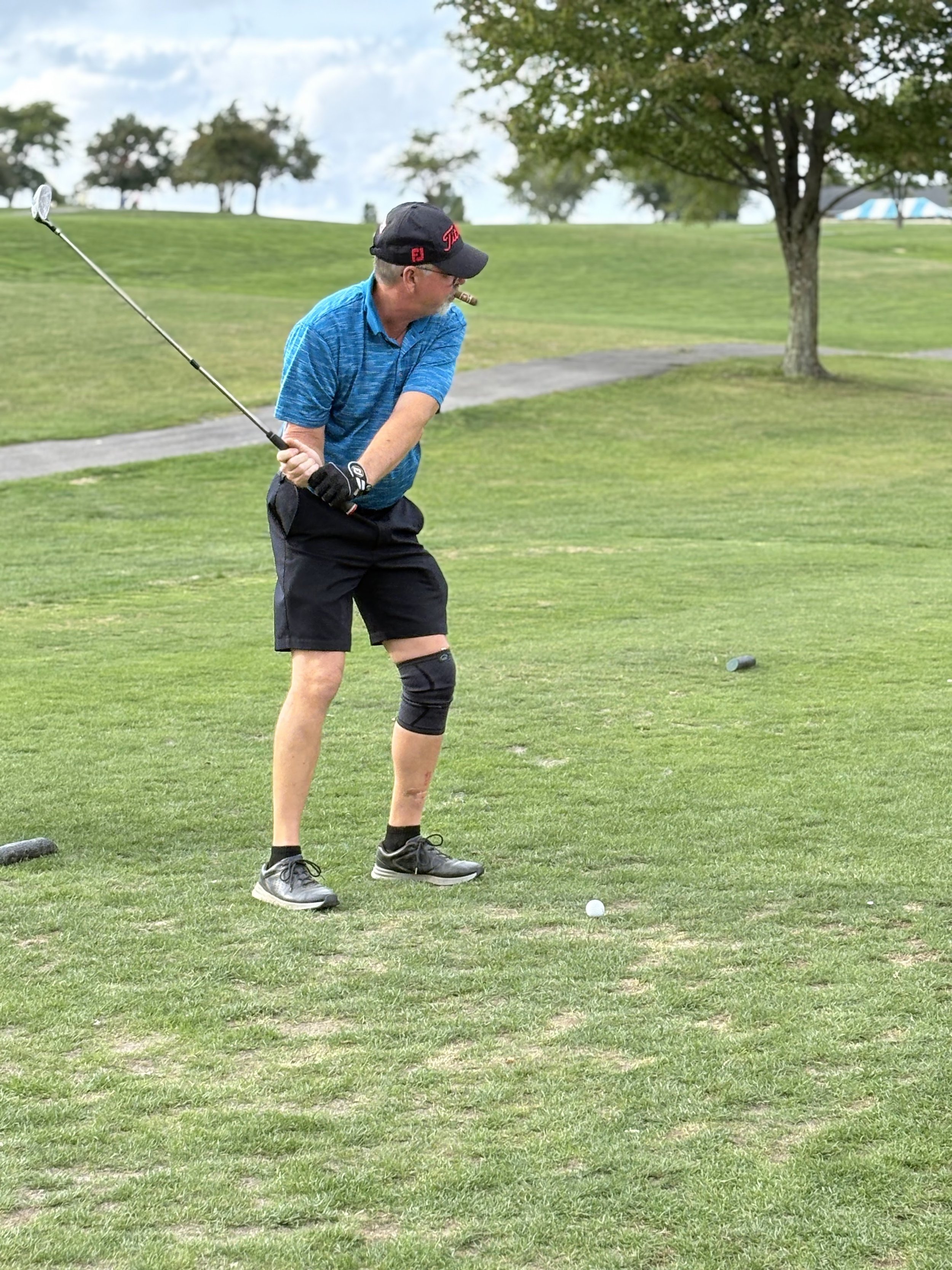 A man wearing a blue shirt, black shorts, and a black cap is playing golf. He is holding a golf club and is in the process of hitting a golf ball on a green golf course with trees and a cloudy sky in the background.