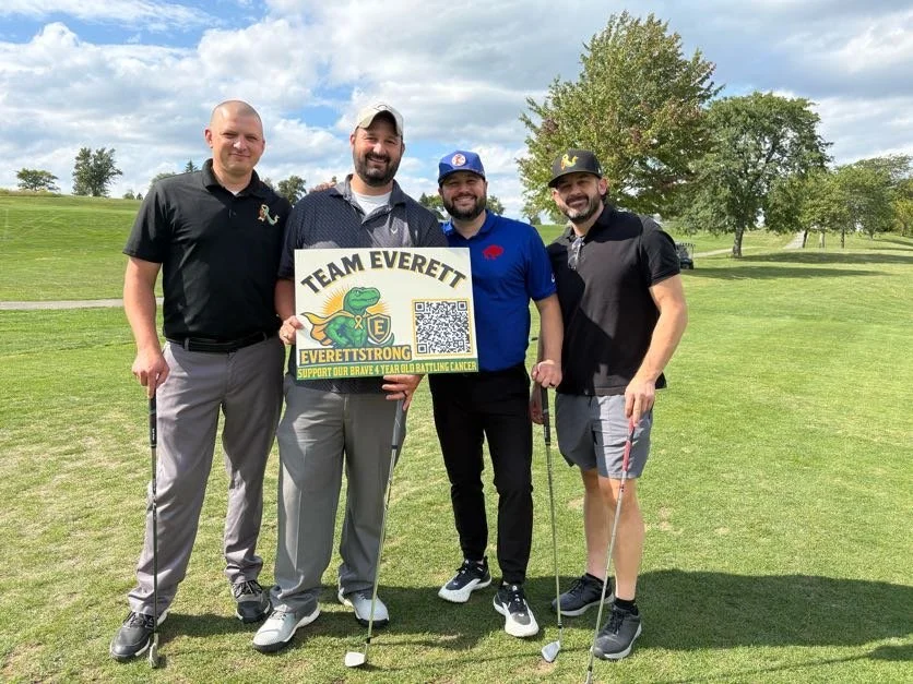 Four men standing on a golf course holding a sign that says "Team Everett, EverettStrong" with a frog mascot, QR code, and message to support childhood cancer battle.