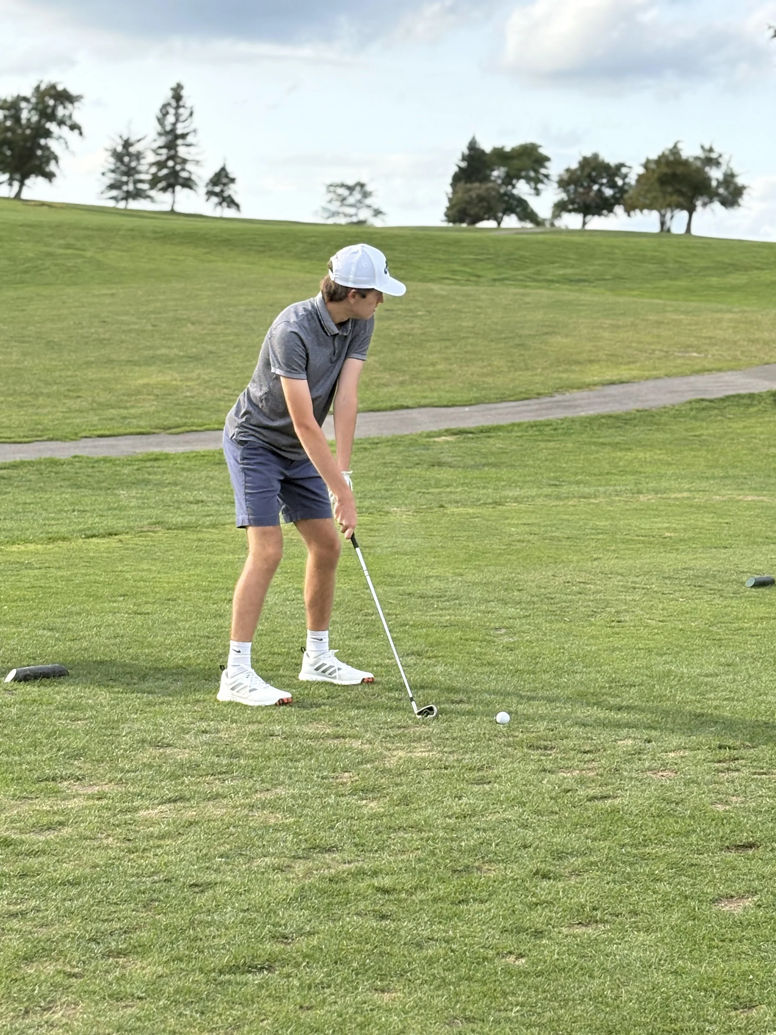 A young man in a grey polo shirt, navy shorts, white sneakers, and a white cap playing golf on a lush green course with a few scattered trees in the background.