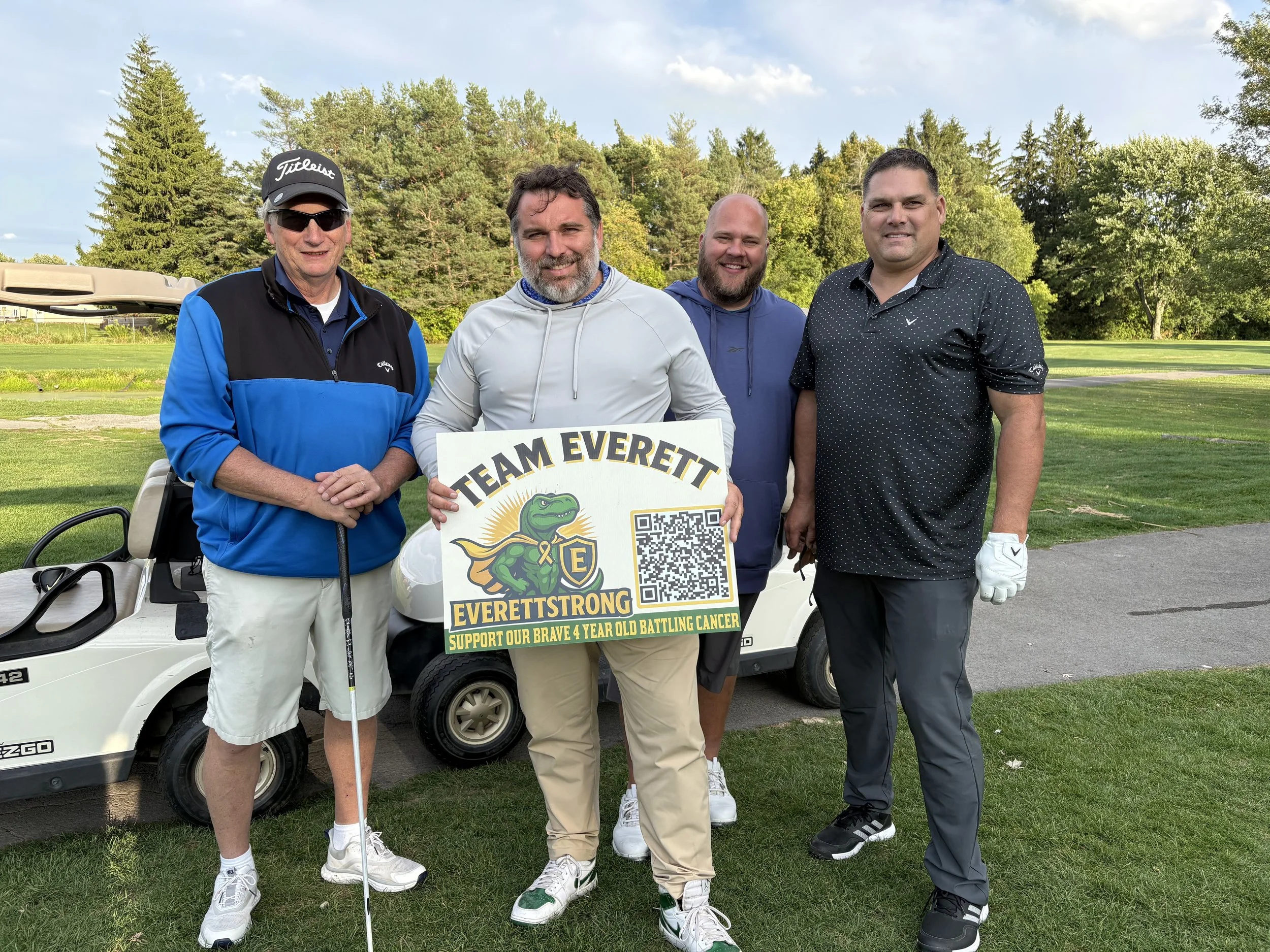 Four men standing together on a golf course, holding a sign that reads 'Team Everett, Everett Strong' with a cartoon alligator logo and a QR code, with trees and a golf cart in the background.