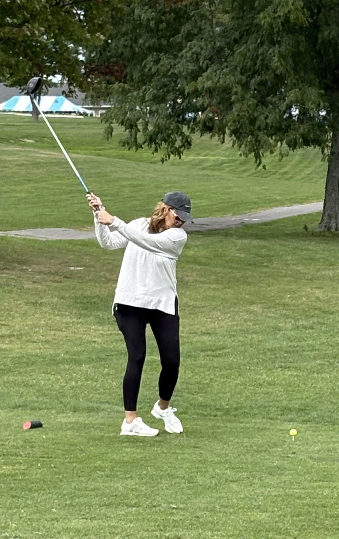 A woman playing golf on a golf course, wearing a white jacket, black pants, white shoes, and a black cap, preparing to hit a golf ball towards a distant hole.