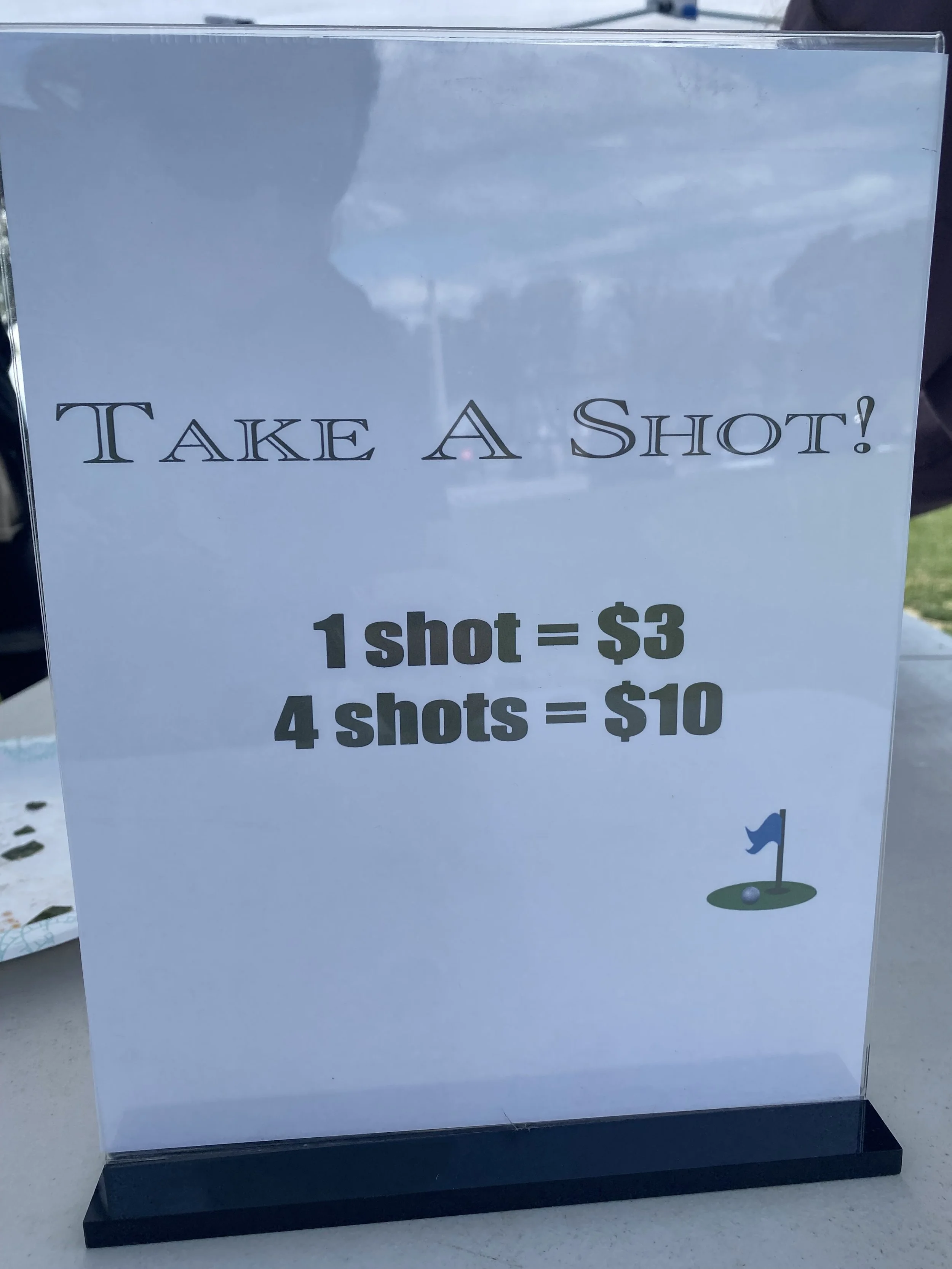 Sign advertising shots for sale, 1 shot for three dollars and 4 shots for ten dollars, with a small graphic of a golf hole and flag.