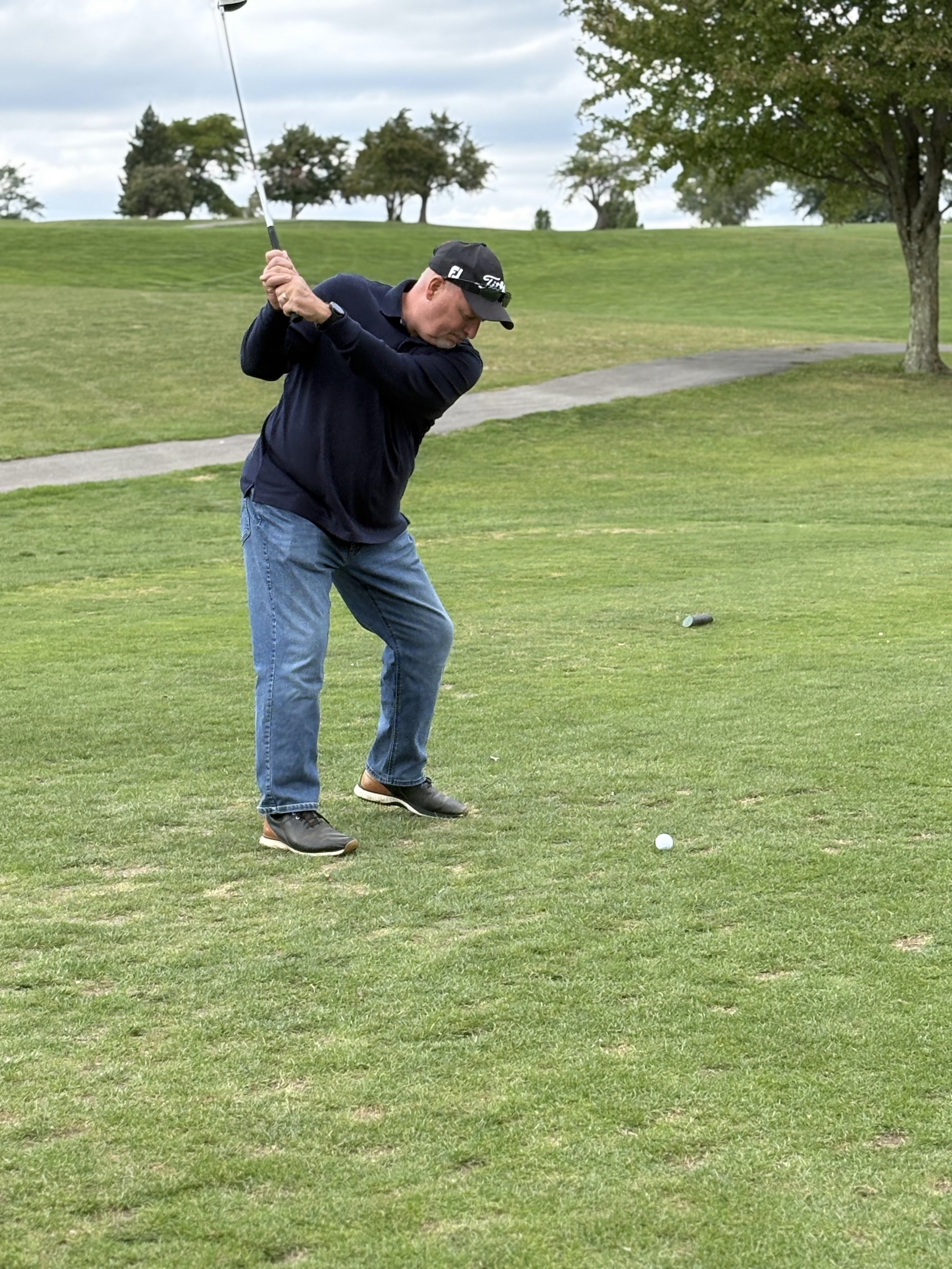 A man in a black cap, black jacket, and jeans playing golf, preparing to hit a golf ball on a golf course with green grass, trees, a walking path, and cloudy sky in the background.